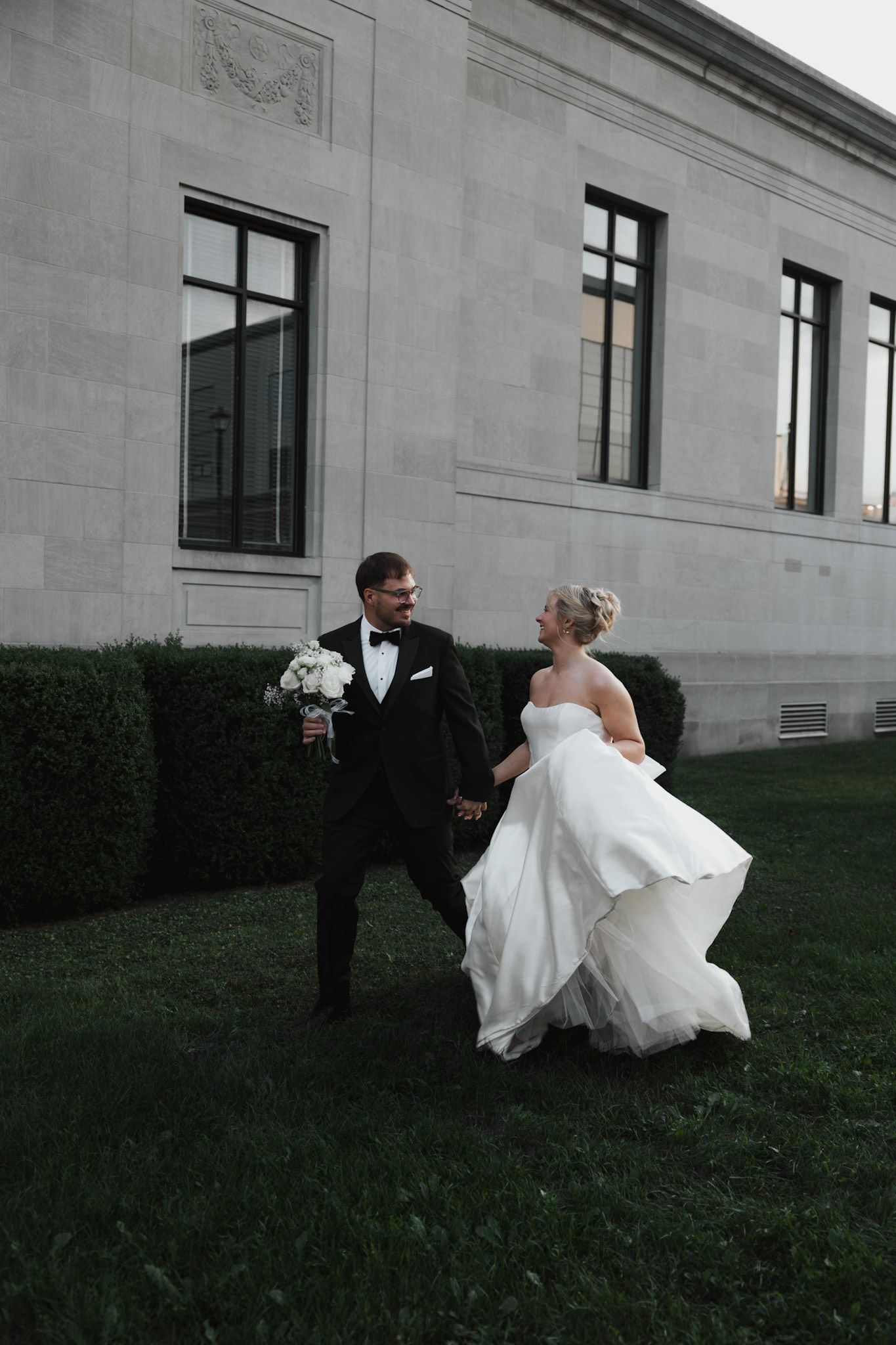 Wedding couple dancing playfully on grass outside a stone building.