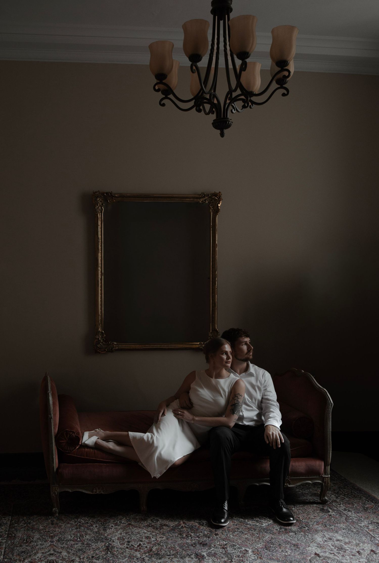 Bride and groom sitting together on a vintage pink sofa beneath a chandelier in a dimly lit elegant room.