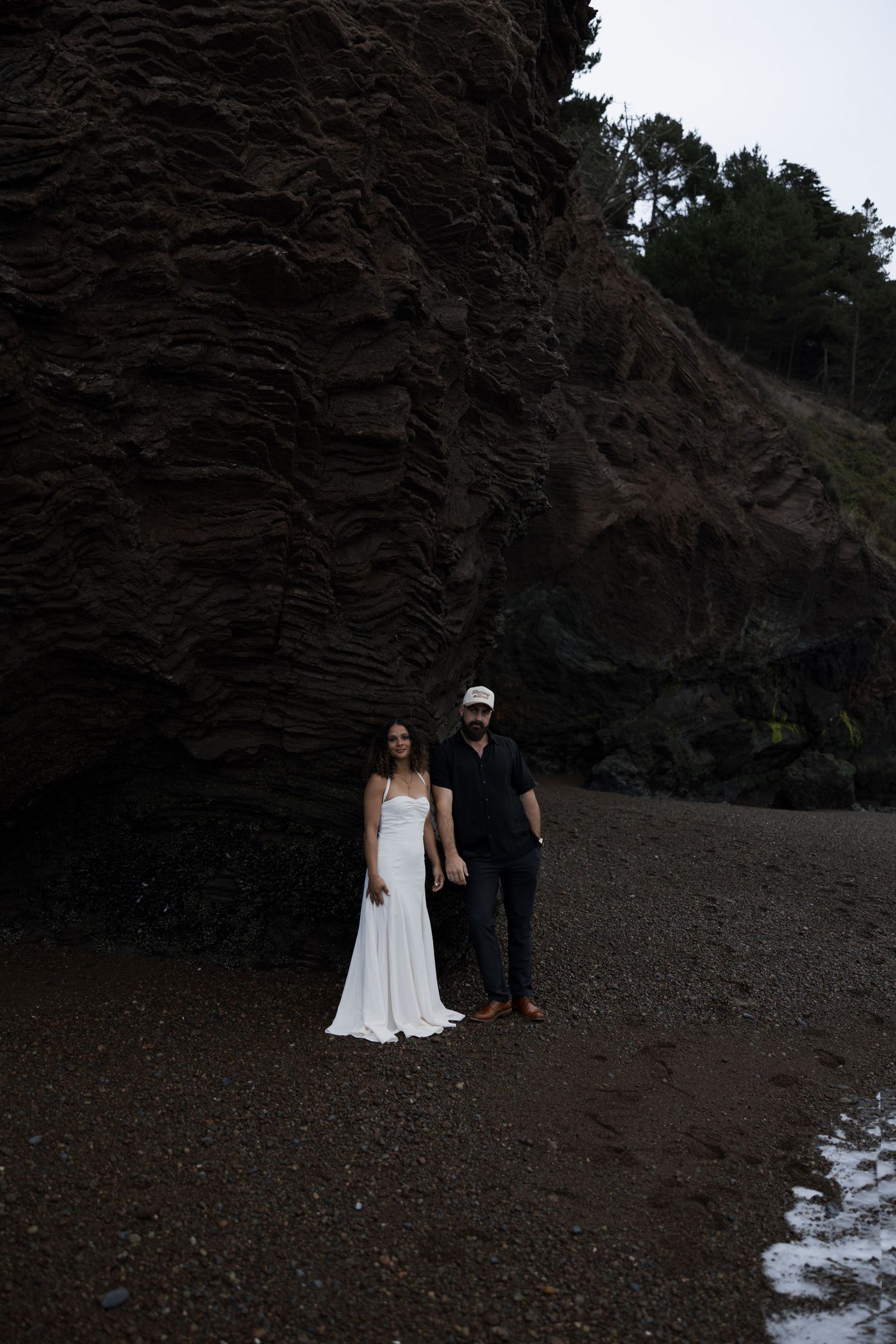 Couple standing against dramatic rocky cliff face on a moody beach.