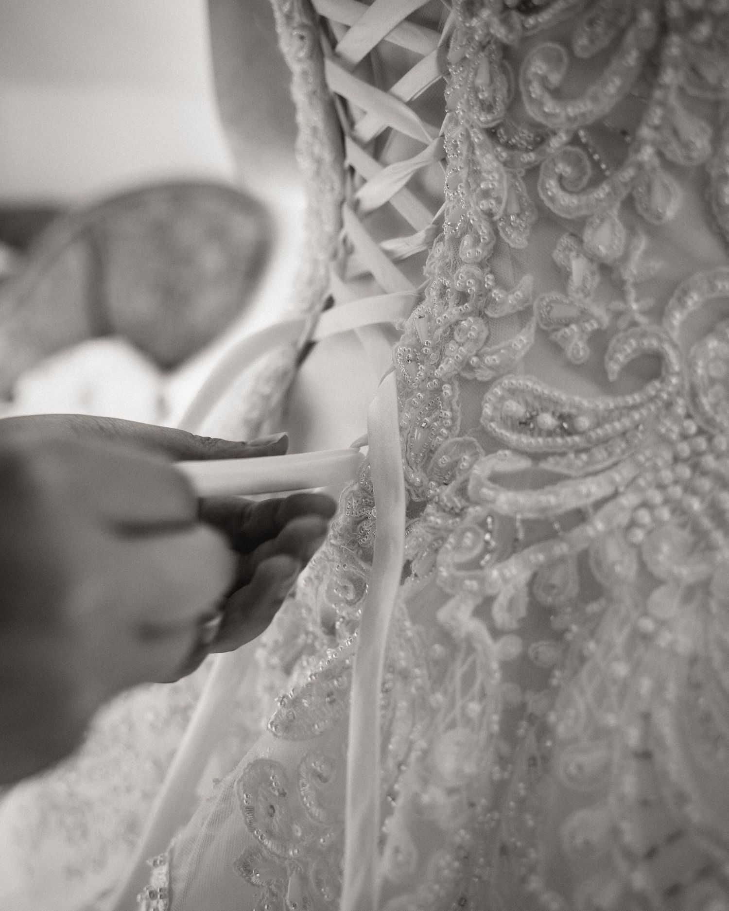 Black and white photo of hands lacing up an ornate beaded wedding dress with intricate embroidery details.