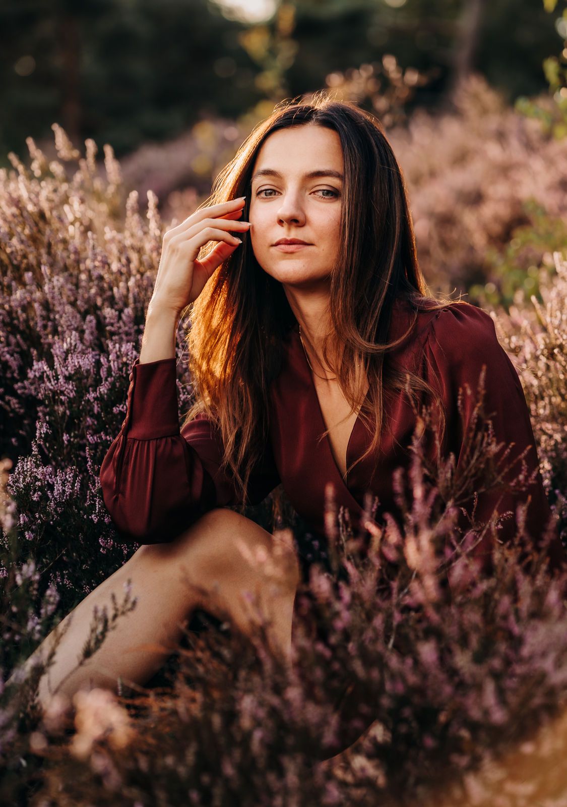 Woman in burgundy dress sitting among purple heather flowers in golden hour light, hand raised to face. Photography in Köln