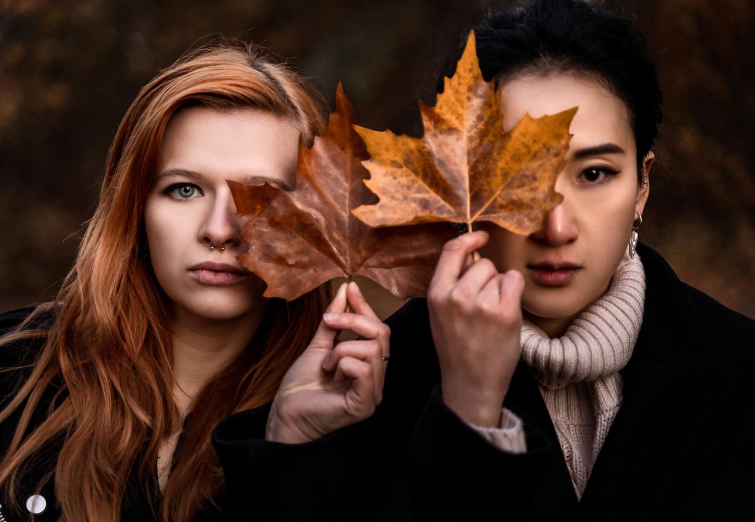 Two women holding autumn maple leaves over their faces in a fall outdoor portrait photography session.