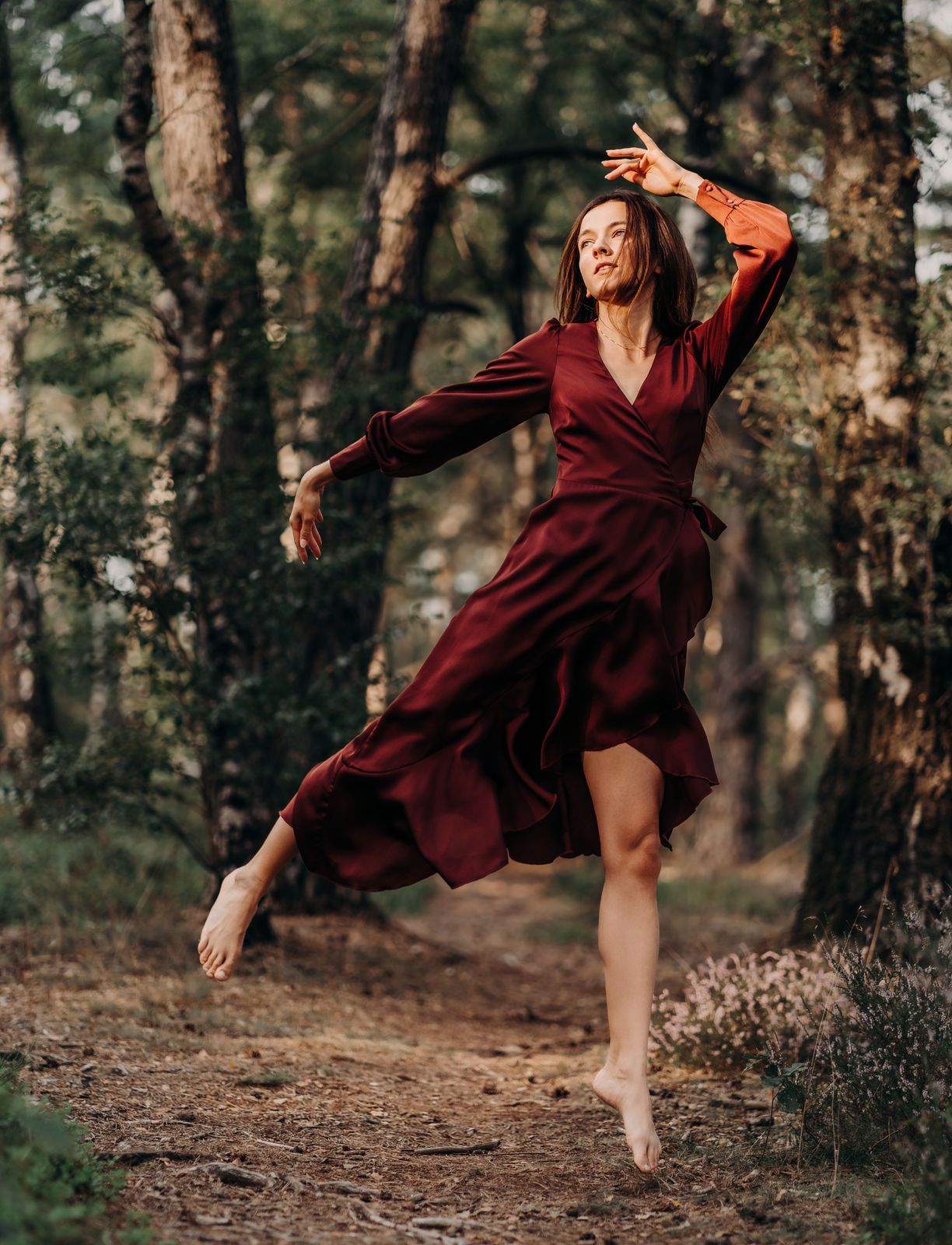 A barefoot woman in a flowing burgundy dress dances gracefully among tall trees in a forest setting.