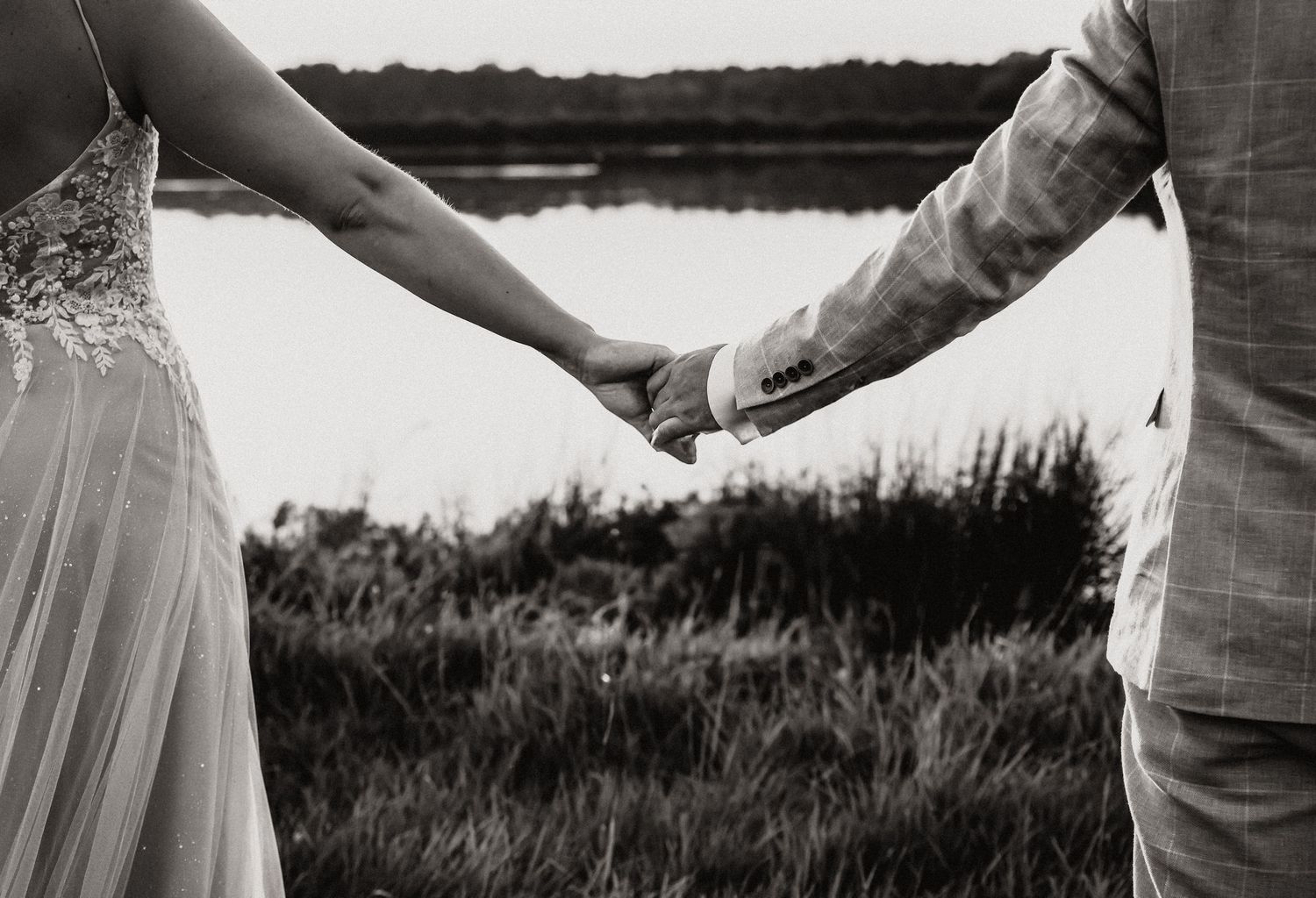 Black and white photo of bride and groom holding hands near a lake surrounded by tall grass.