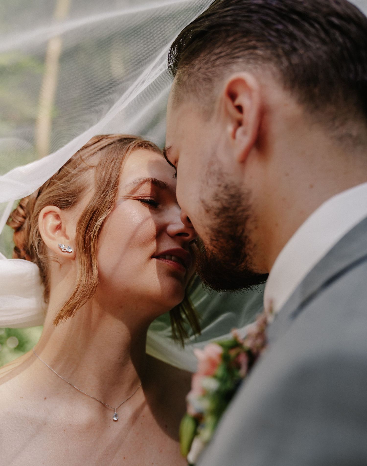 Braut und Bräutigam teilen sich einen zärtlichen Moment unter einem Hochzeitsschleier, wobei der Bräutigam die Stirn der Braut sanft küsst.