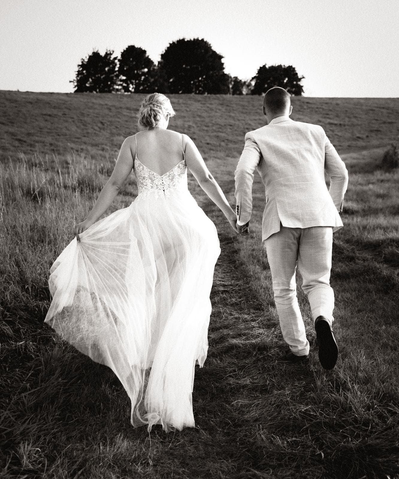 Schwarz-Weiß-Foto von Braut und Bräutigam, die sich an den Händen halten und an ihrem Hochzeitstag durch ein Feld laufen. Alfshausen
