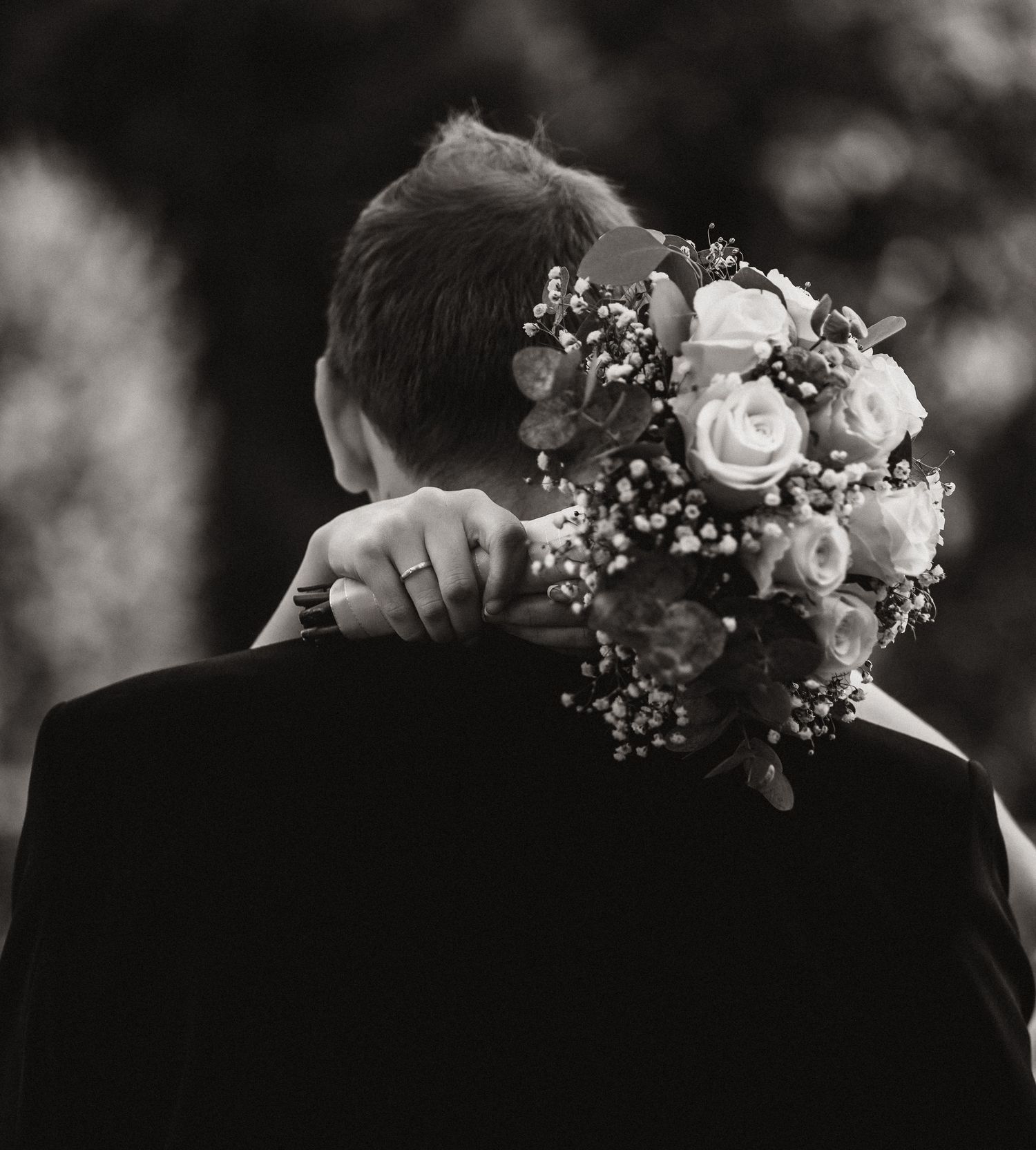Black and white photo of a couple embracing, bride holding a rose bouquet over groom's shoulder at a wedding.