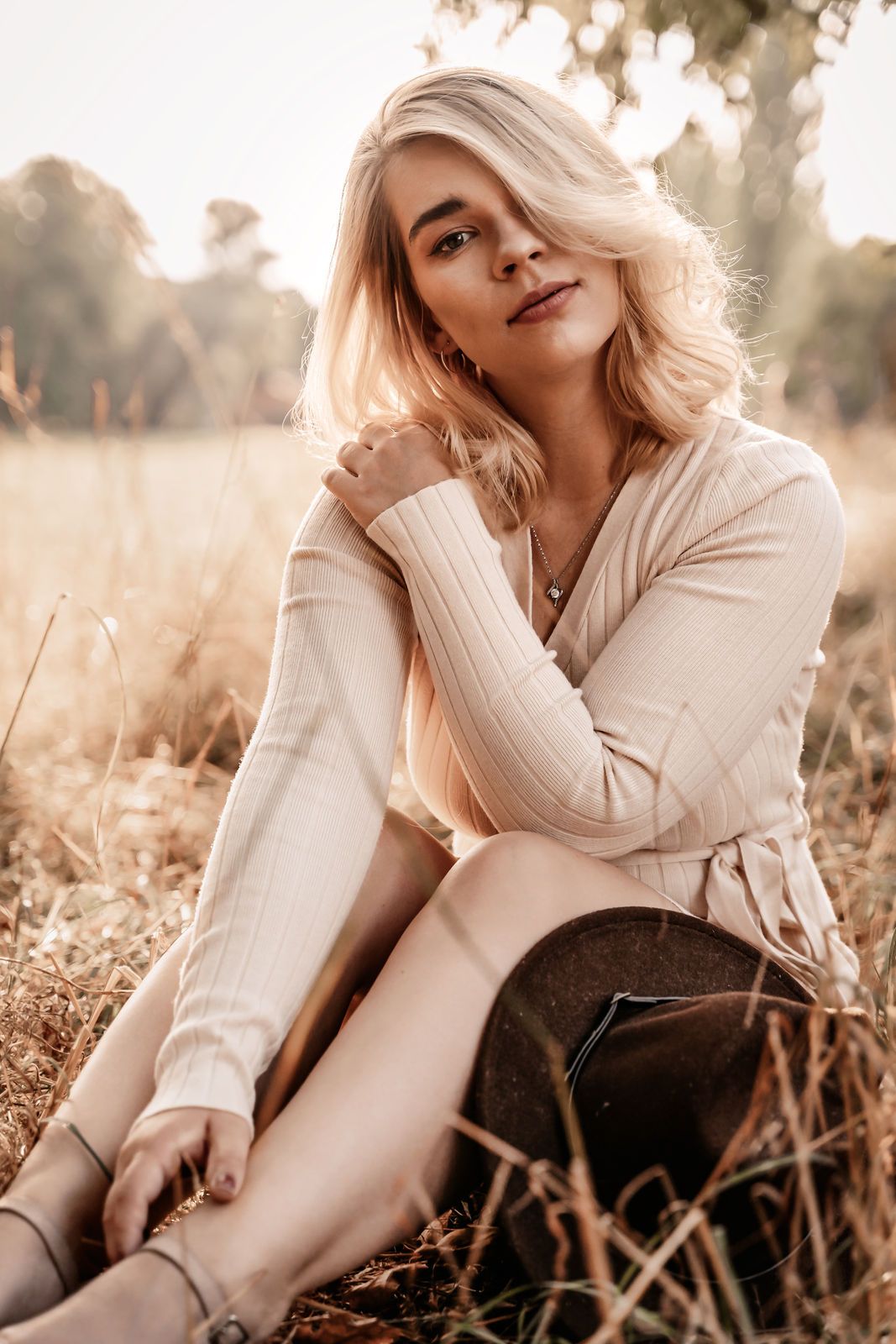 Blonde woman in cream ribbed sweater sitting in golden field grass, smiling softly at camera on sunny autumn day. Bad Iburg