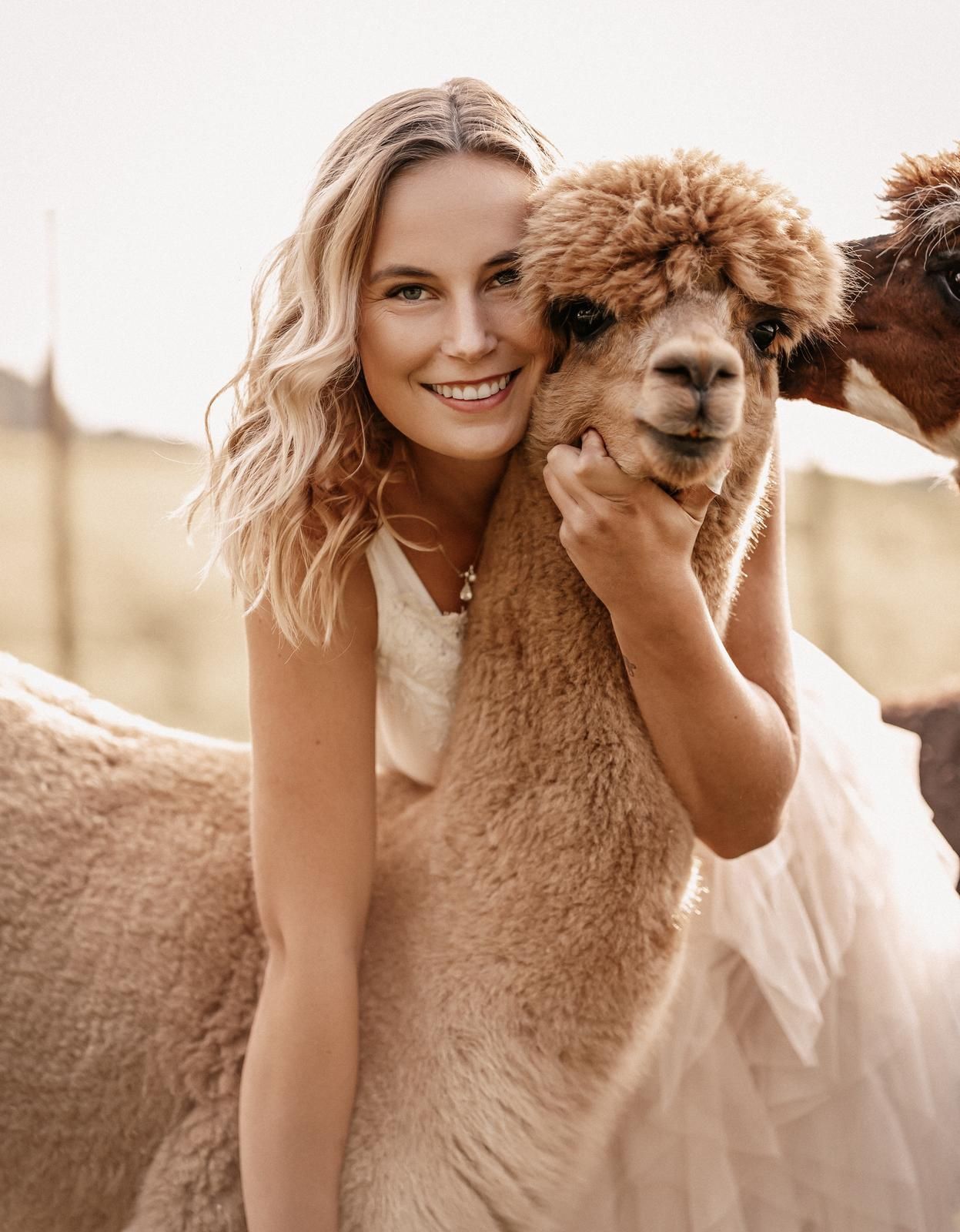 Smiling blonde woman in white dress hugging a fluffy brown alpaca outdoors on a farm.