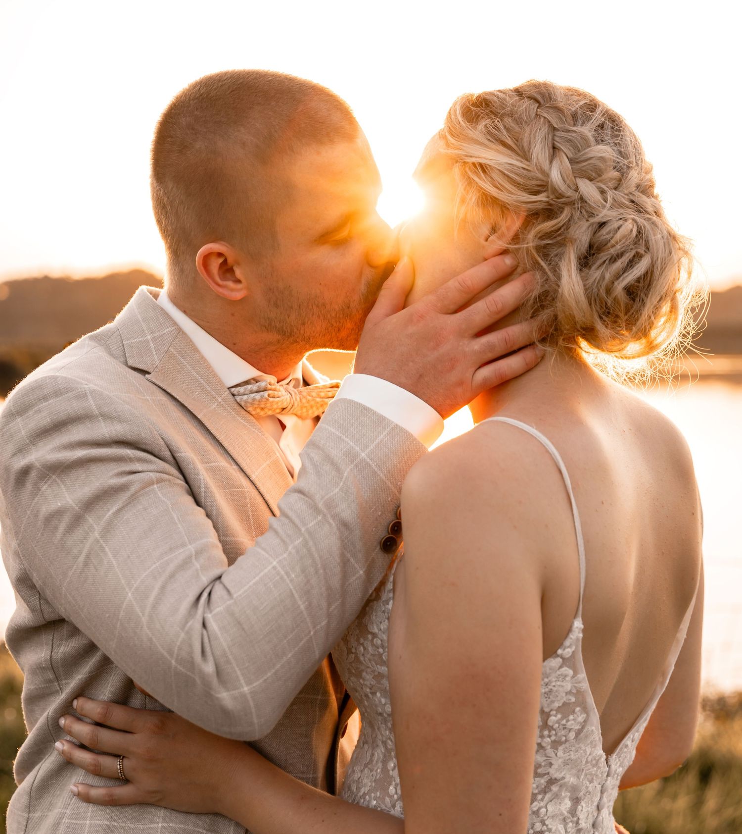 Bride and groom sharing a romantic sunset kiss, groom in plaid suit holding bride in lace wedding dress.