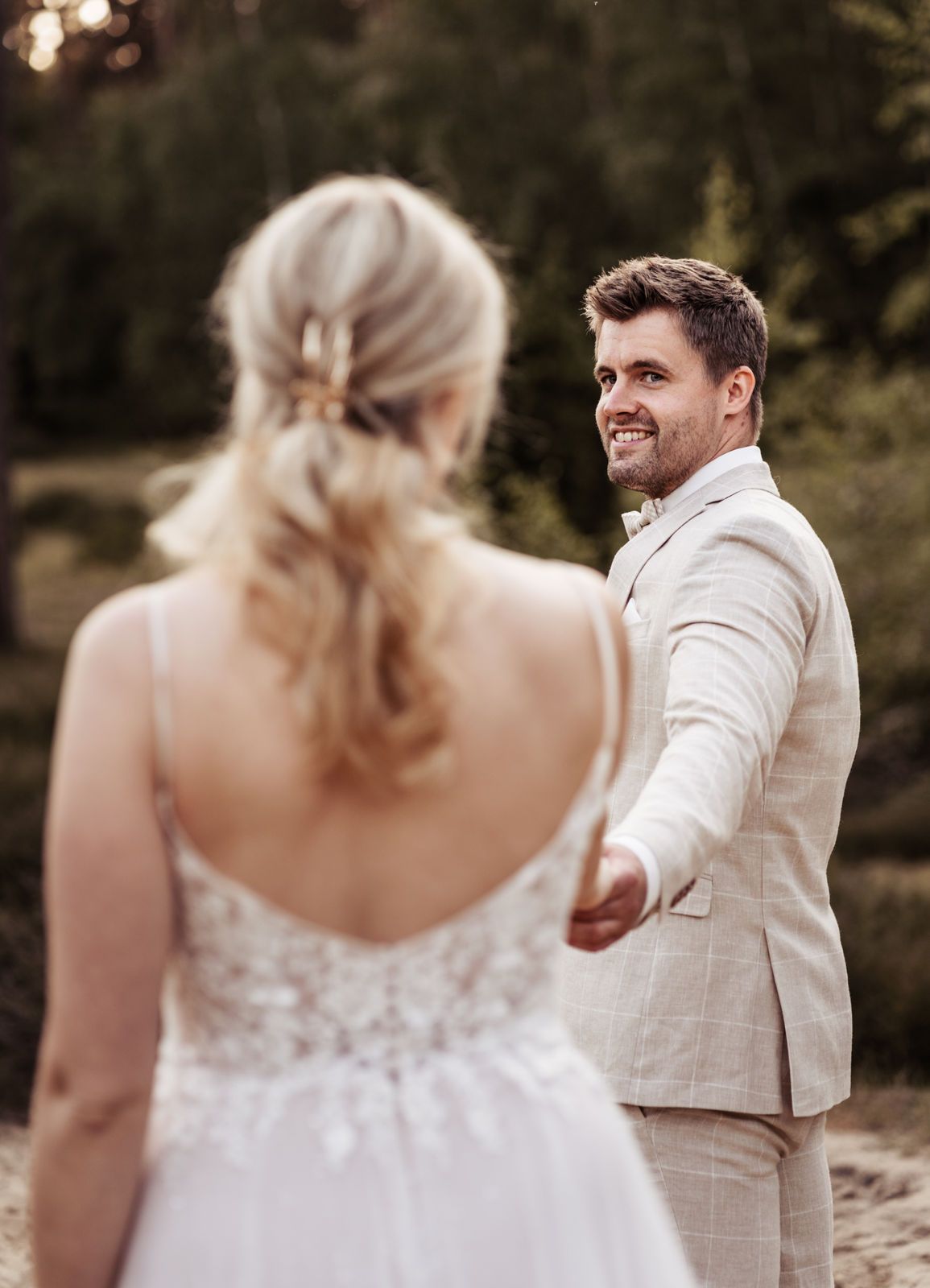 Ein lächelnder Bräutigam in einem beigen Anzug greift während eines romantischen Hochzeitsmoments im Freien nach der Hand seiner Braut.