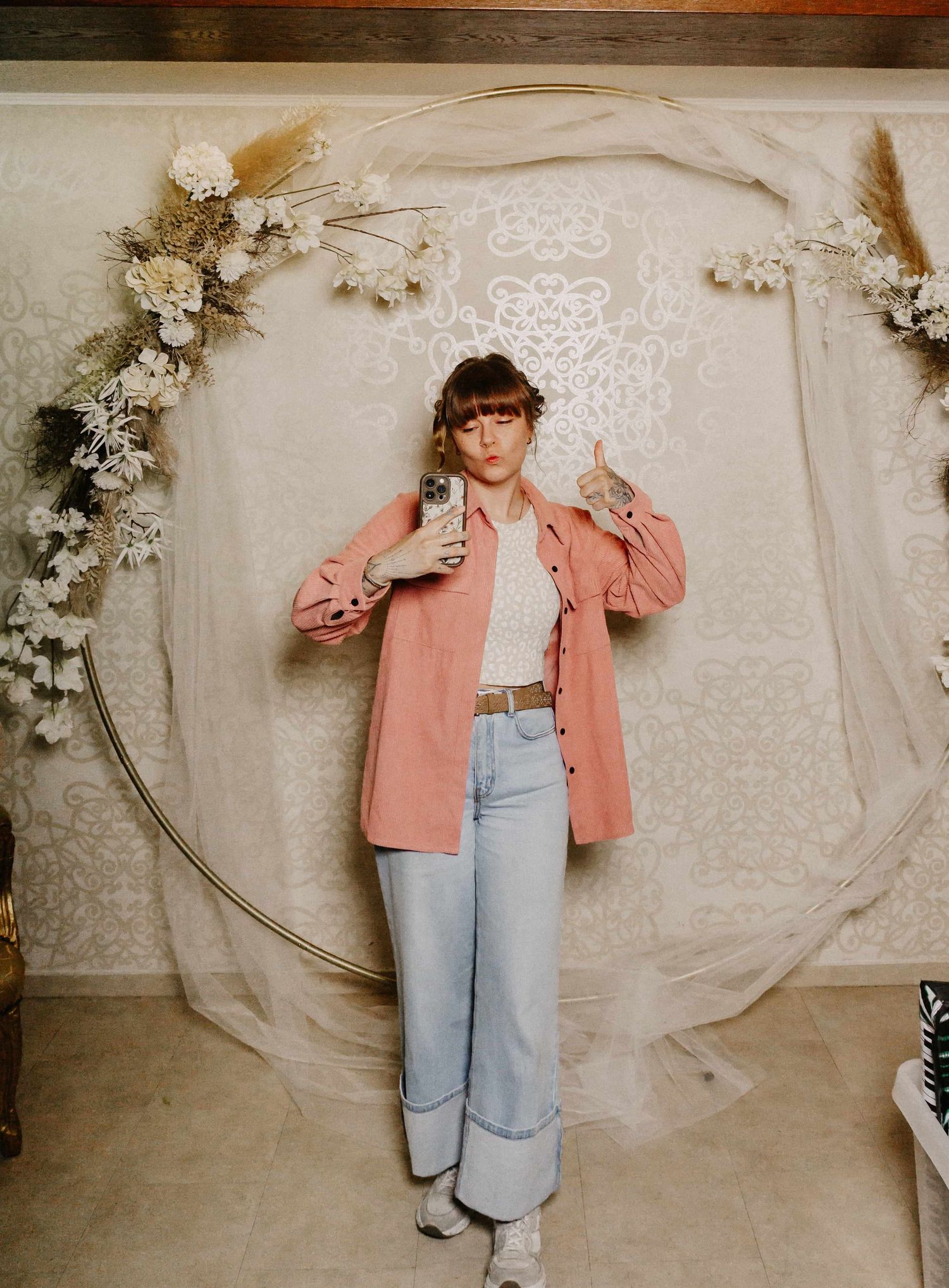Person in pink blazer and wide-leg jeans poses for mirror selfie in front of floral hoop backdrop.