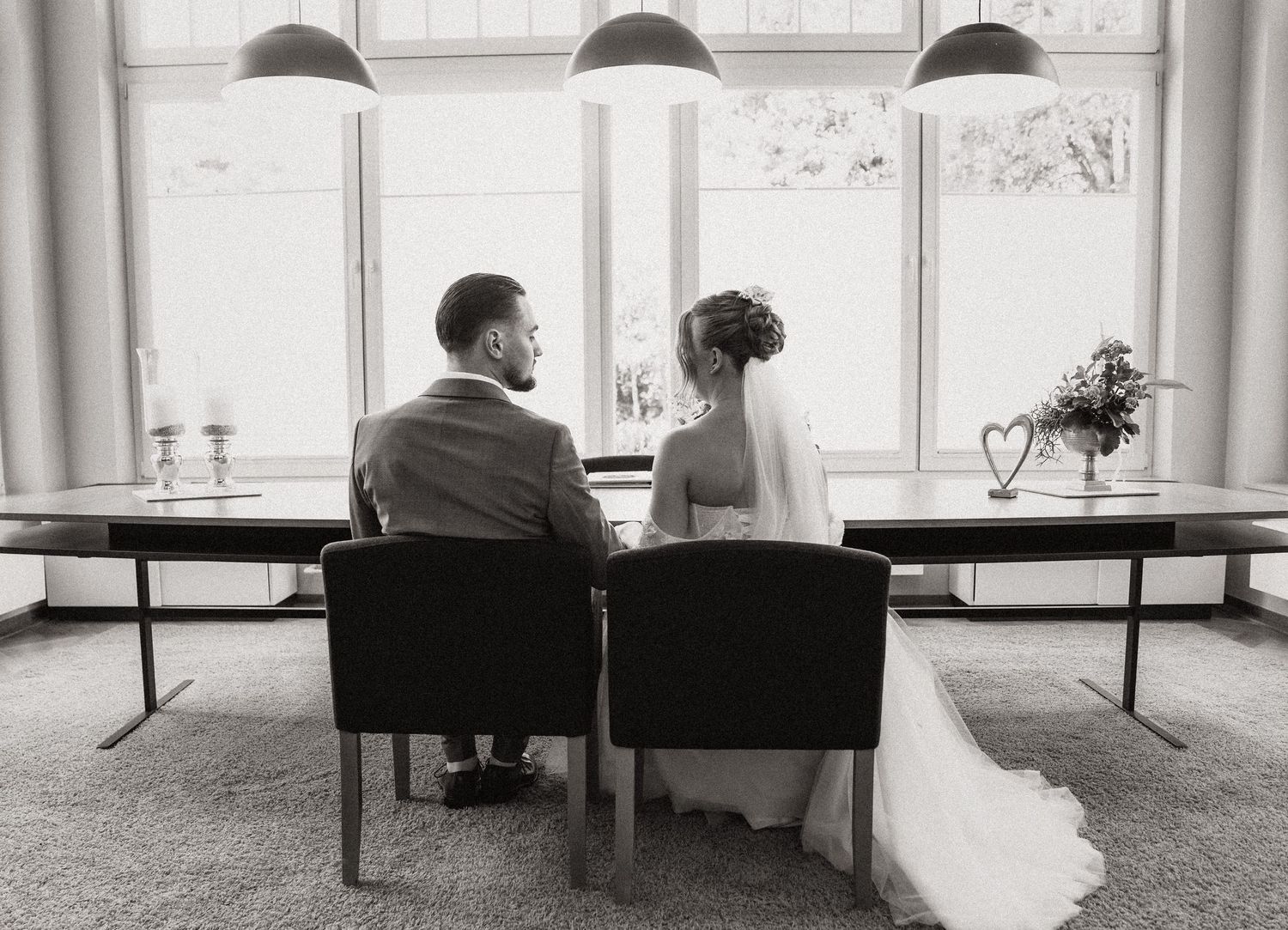 Schwarz-Weiß-Foto von Braut und Bräutigam, die an ihrem Hochzeitstag zusammen an einem Tisch sitzen und einem großen Fenster zugewandt sind.