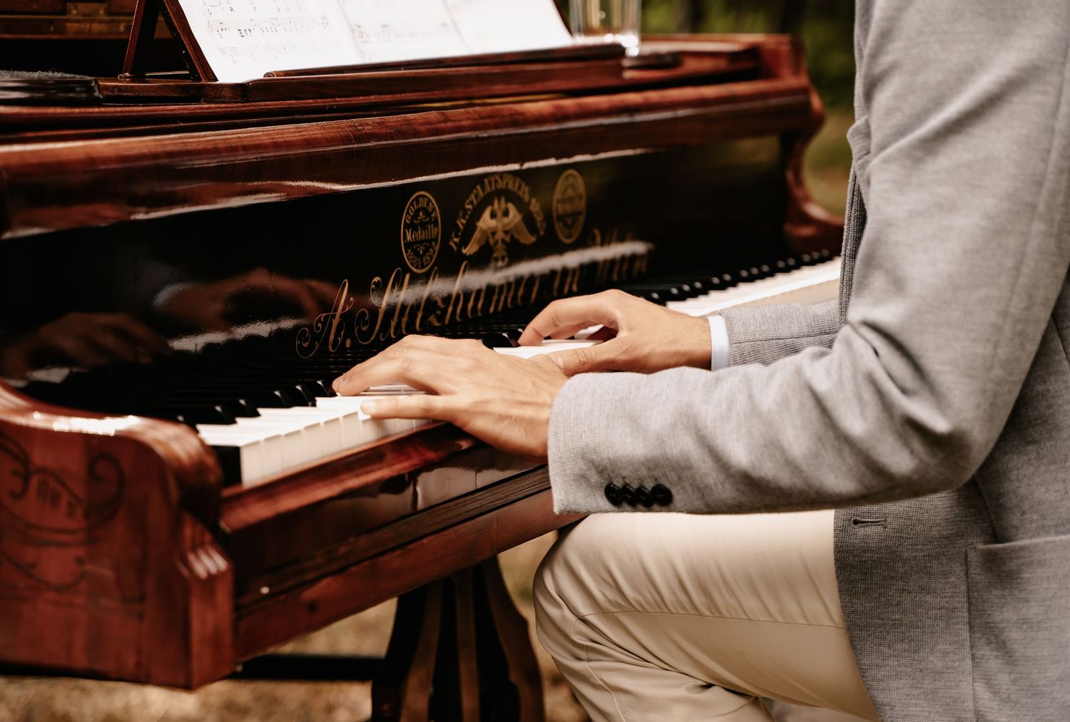 Man in gray suit playing a vintage mahogany upright piano outdoors, hands on keys in elegant performance.
