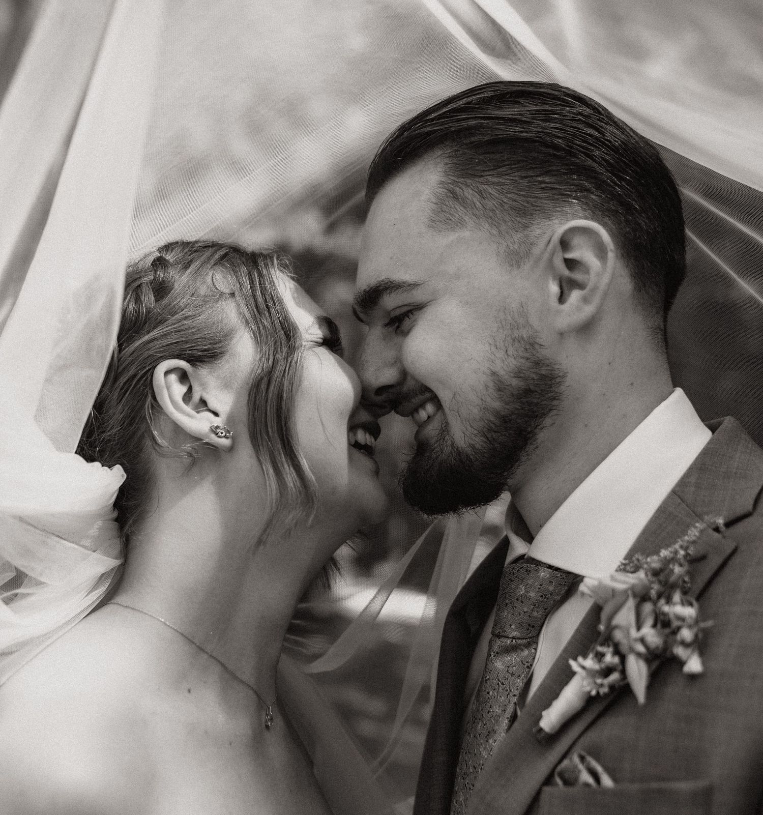 Schwarz-Weiß-Foto einer lächelnden Braut und eines Bräutigams, die sich unter einem Hochzeitsschleier die Stirn berühren.