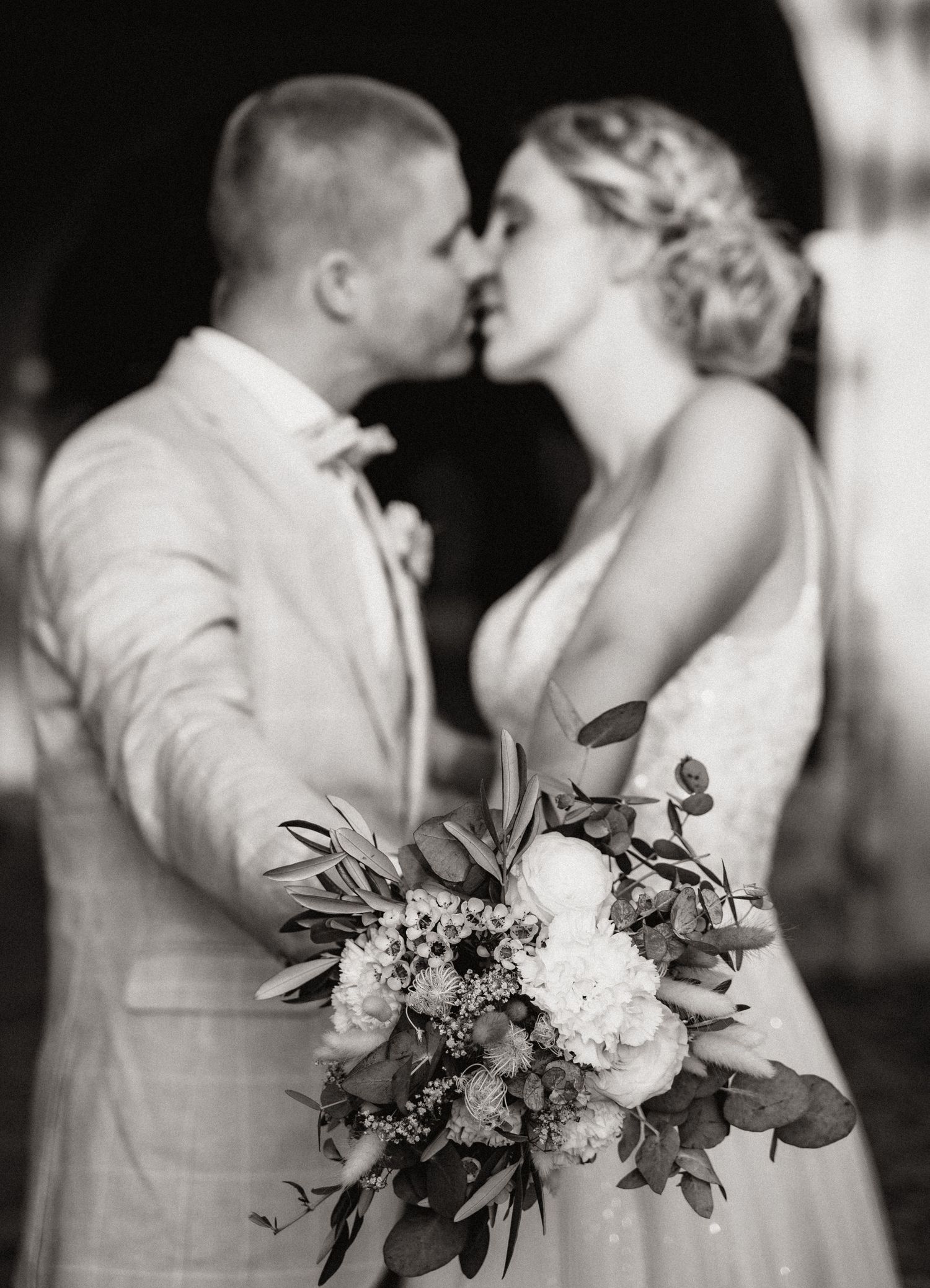 Black and white wedding photo of bride and groom kissing, bride holding lush floral bouquet with roses and eucalyptus.