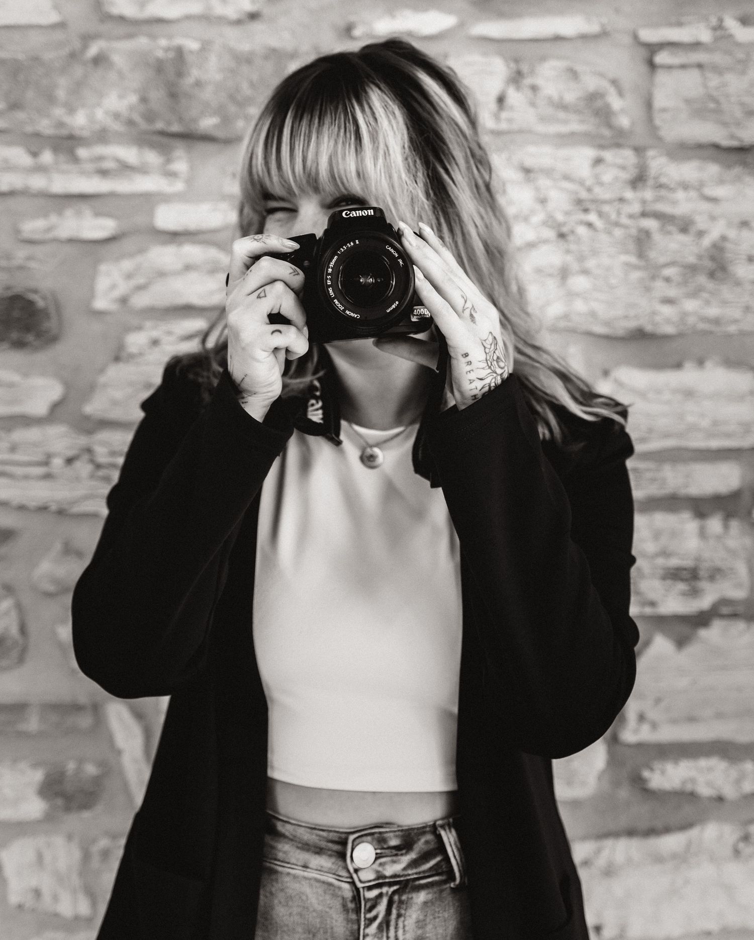 Black and white photo of a woman holding a Canon camera up to her face, wearing a black blazer and light top.
