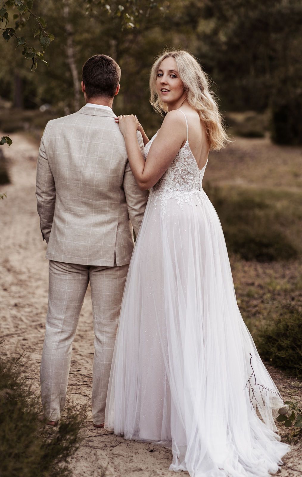 Bride in white lace spaghetti strap gown looks back while holding groom's arm in beige suit outdoors. Muenster und Telgte. Hochzeitsfotograf