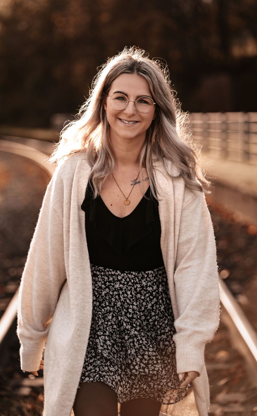 Young woman with glasses and blonde hair smiles on railroad tracks, wearing floral skirt and cream cardigan.