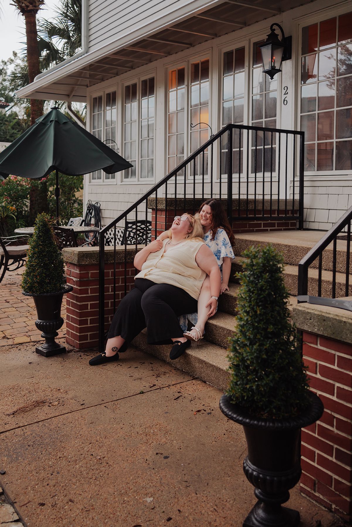 Two women laughing and relaxing on brick steps outside a charming white building with decorative topiaries.