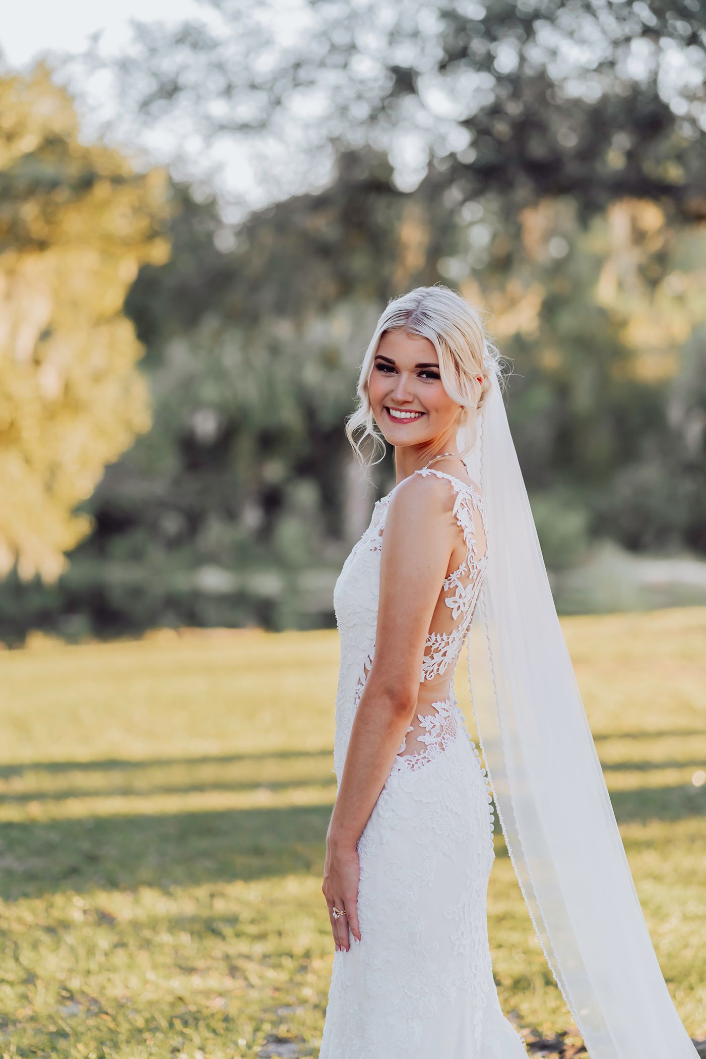 A bride in a white lace wedding dress with a flowing veil poses outdoors in a sunlit field for wedding photographer in Ocala Florida.