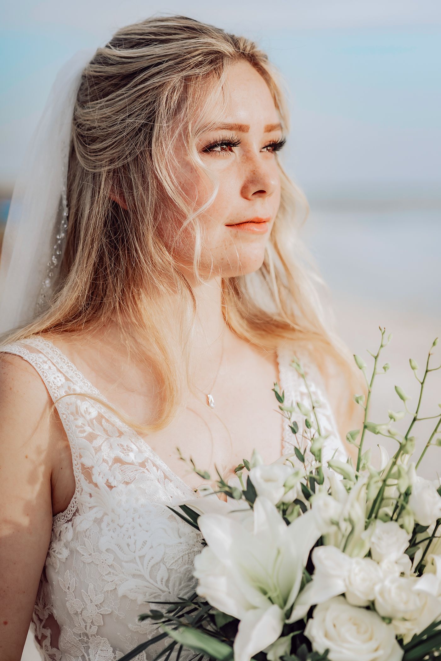 A bride in a white lace dress holds a stunning bouquet of white flowers and delicate greenery against a soft background.