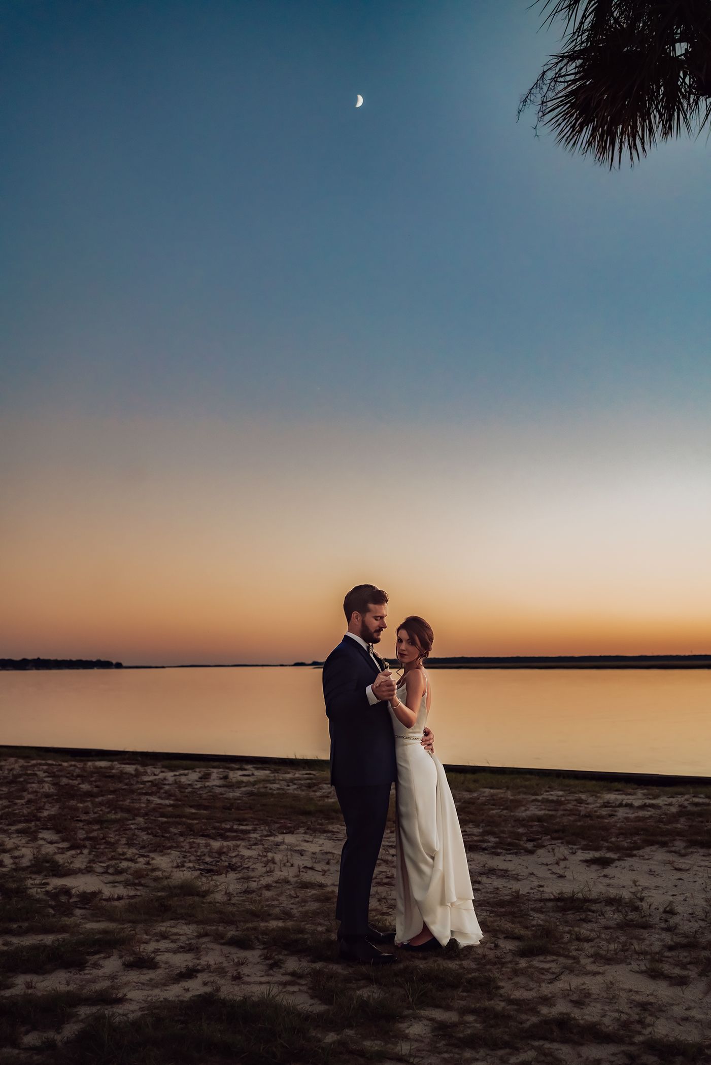 A couple shares a romantic moment at sunset on a beach with a warm orange sky and dark sand.