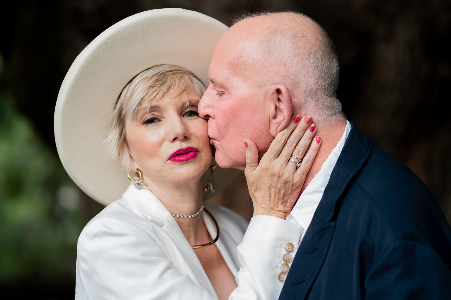 Romantic couple in formal attire share an intimate moment outdoors, with one wearing a stylish white hat and bright lipstick.