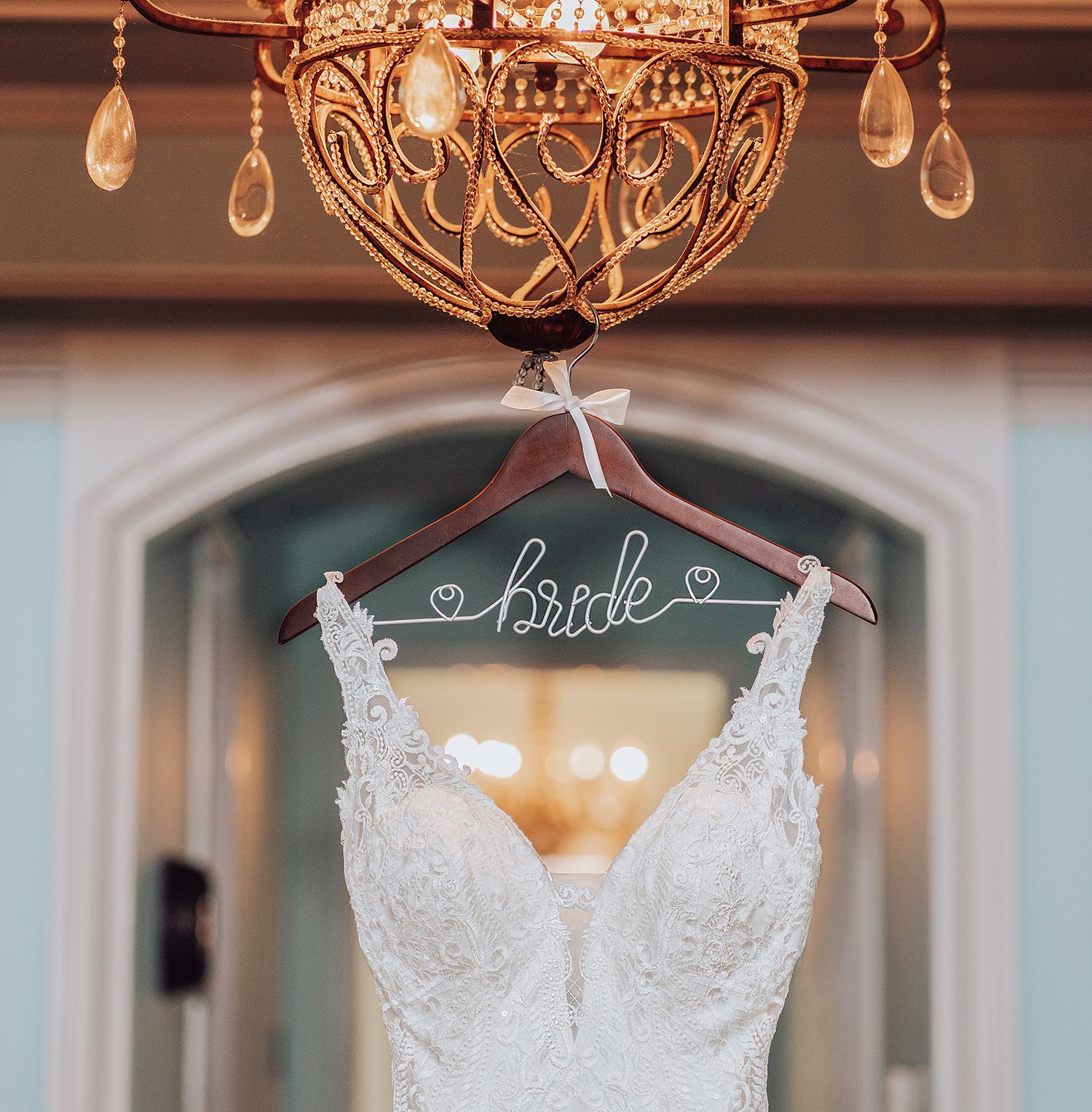 Elegant white lace wedding dress with beaded details hanging from crystal chandelier in luxurious venue.