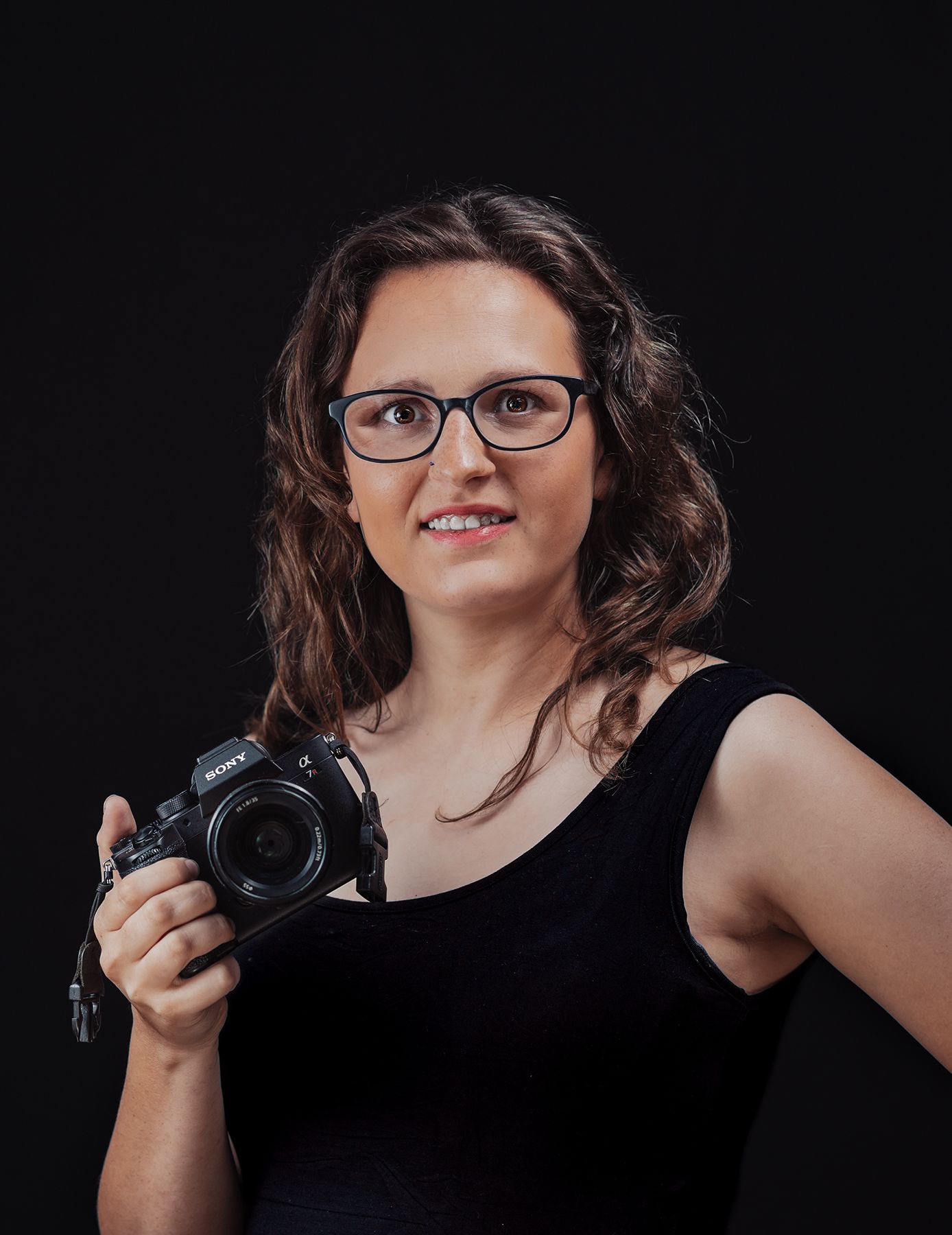 Professional photographer wearing glasses and a black sleeveless top holds a DSLR camera against dark background.