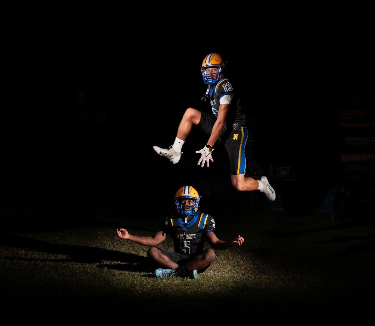 Two football players in blue and gold uniforms pose dramatically at night, one jumping over the other who sits on the field.