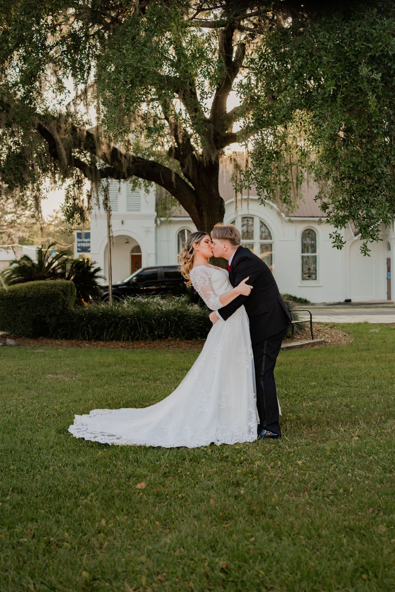 Bride and groom share a romantic kiss under a moss-draped tree on a green lawn outside a white wedding venue.