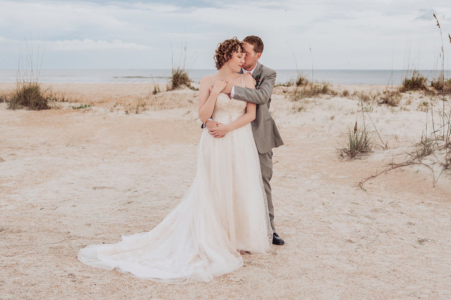 A couple in wedding attire embraces on a sandy beach with sea grass and cloudy skies in the background.