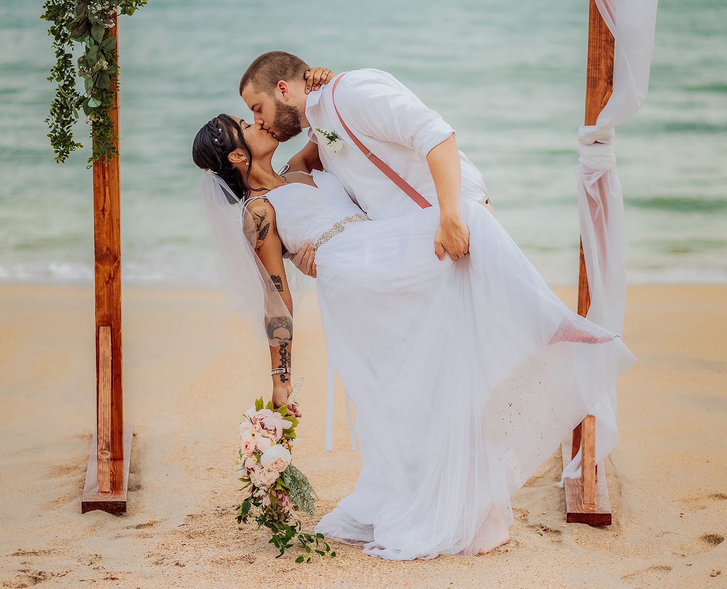 A romantic beach wedding ceremony with a couple sharing a kiss under a floral arch by the ocean.