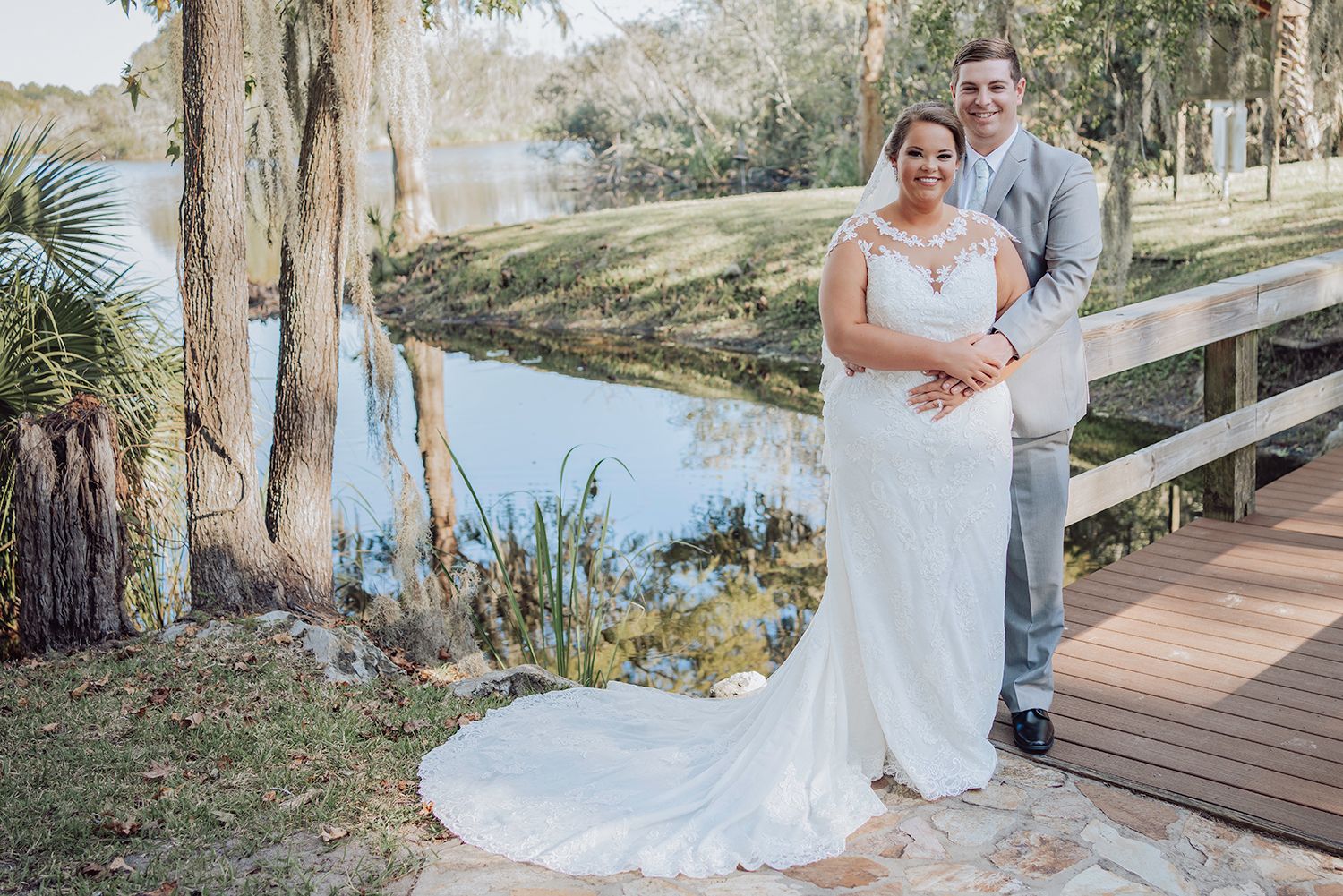 A romantic wedding photoshoot on a wooden boardwalk surrounded by lush trees and a serene lake in the background.