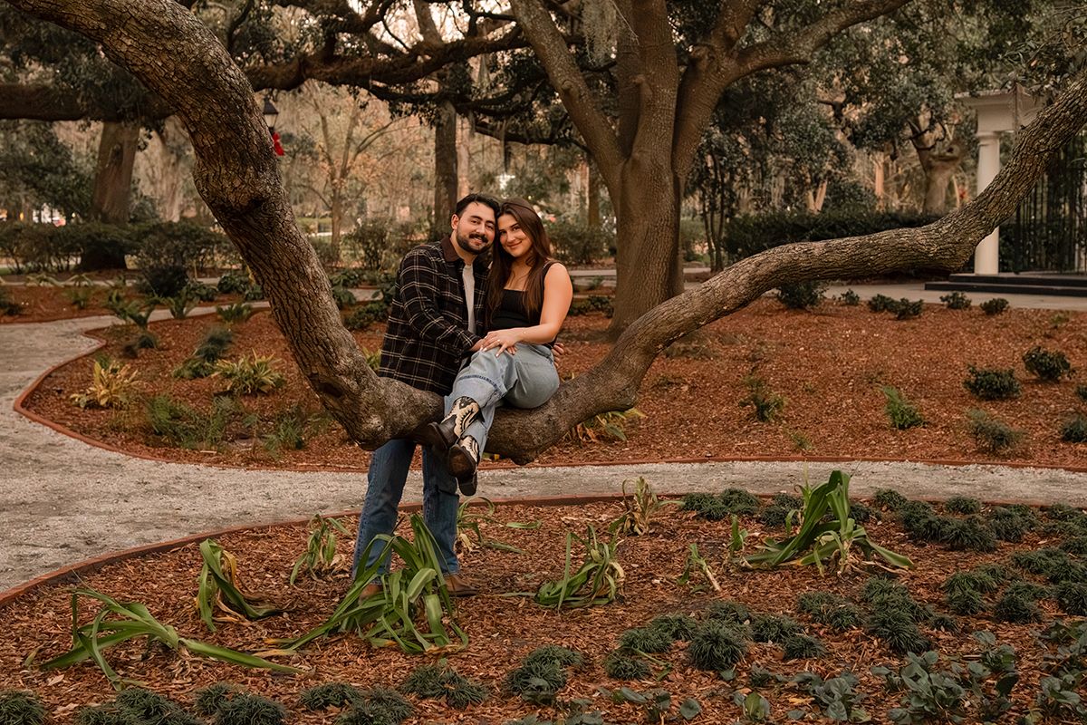 Couple smiling and posing together on a large tree branch in a scenic park with lush greenery.