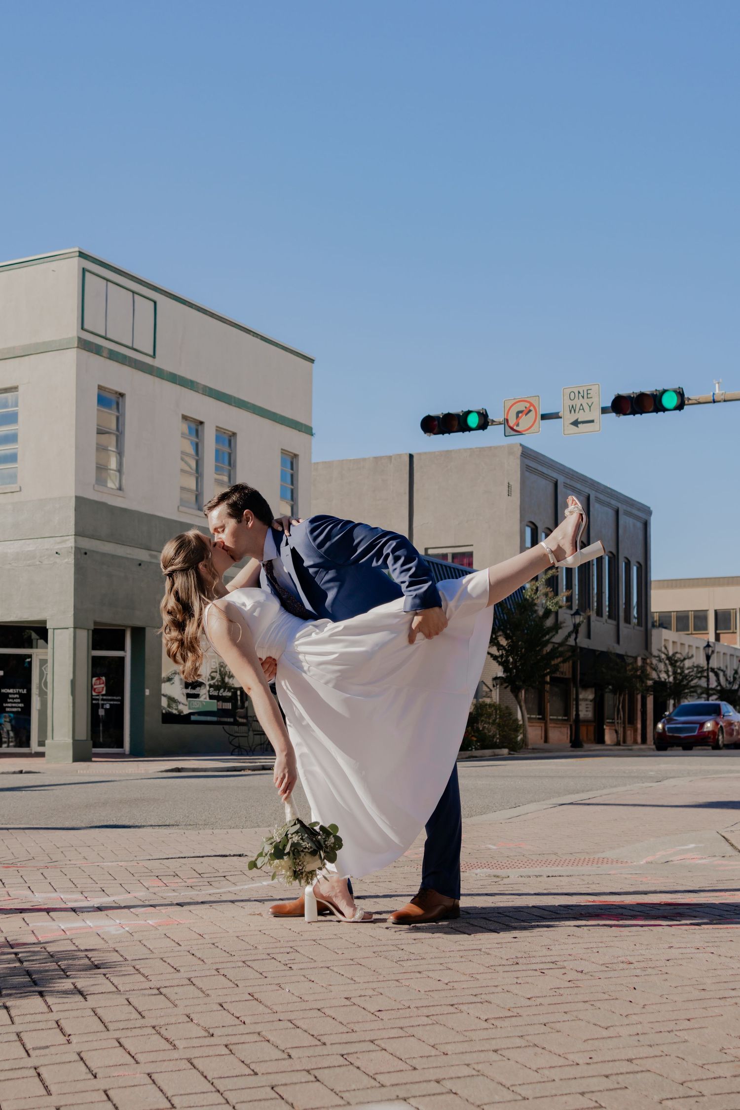 Groom lifts bride in white dress at a sunny urban intersection, sharing a romantic kiss on their wedding day.