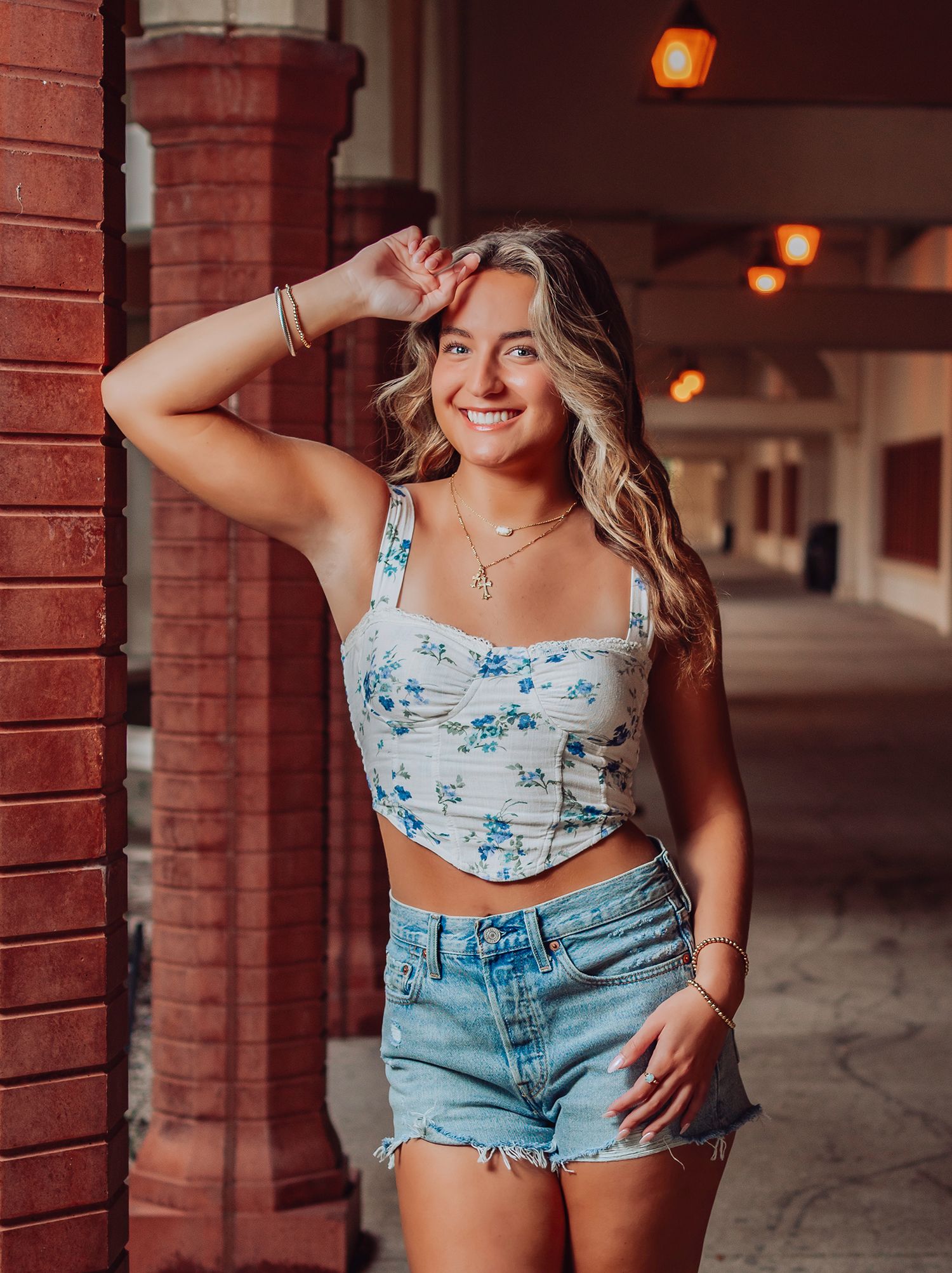 High school senior in a floral crop top and denim shorts poses against a brick wall with warm lighting in an urban setting in St. Augustine