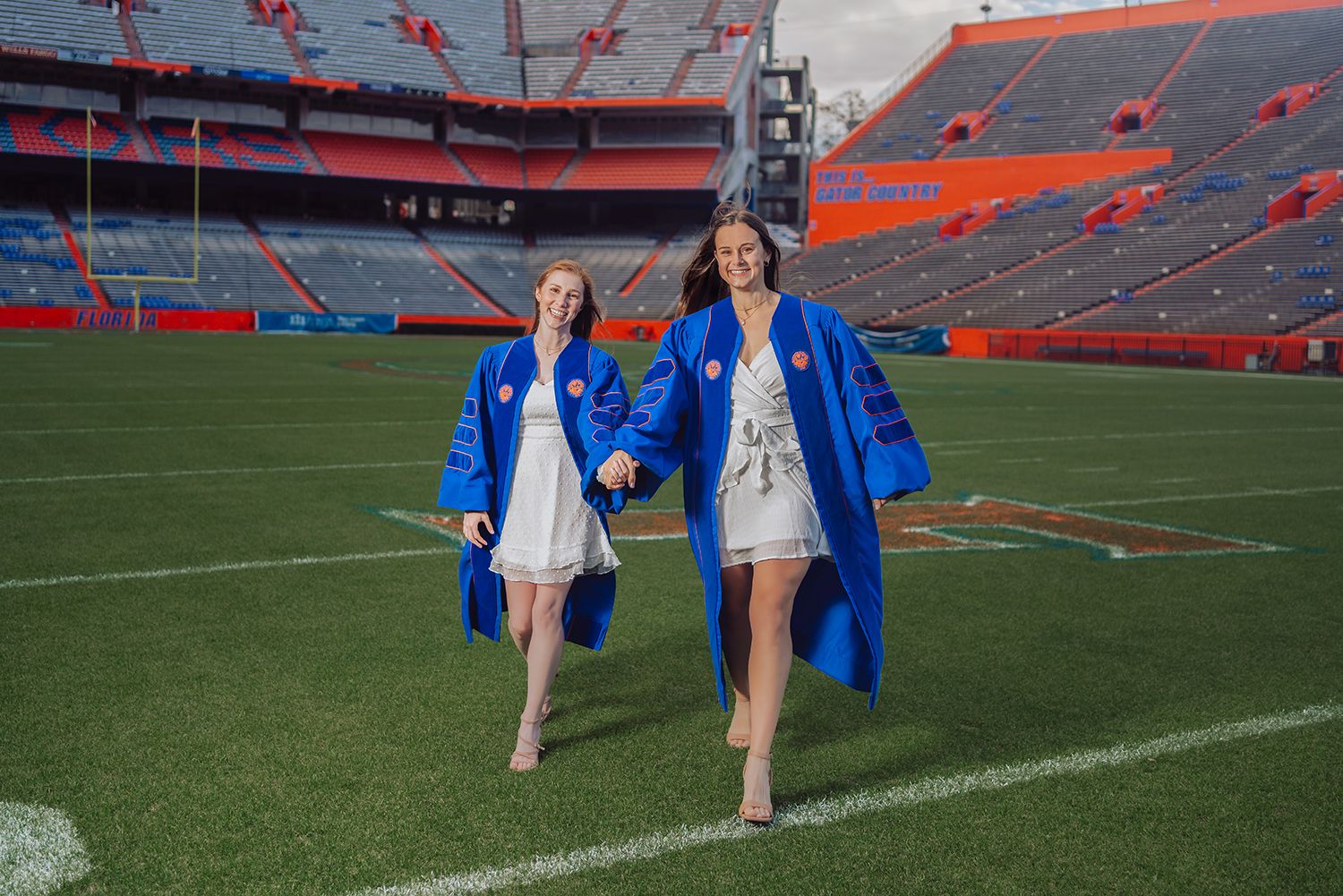 Two graduates in blue robes walk across the field at Ben Hill Griffin Stadium at the University of Florida in Gainesville, Florida.