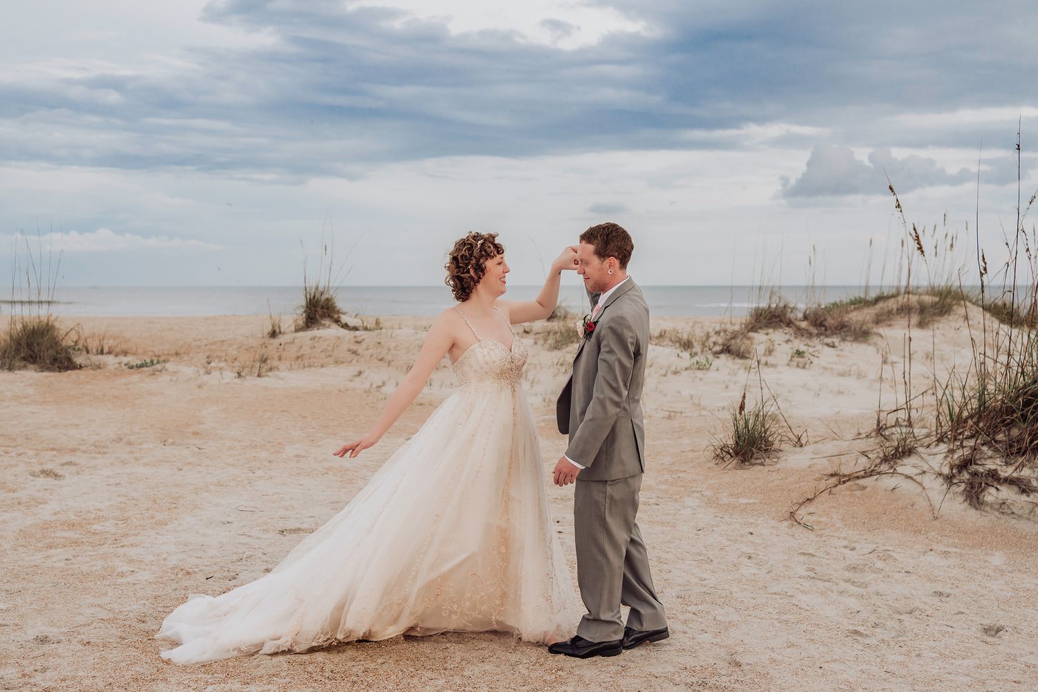 A couple walks along a sandy beach with dunes and sea grass under a cloudy sky.