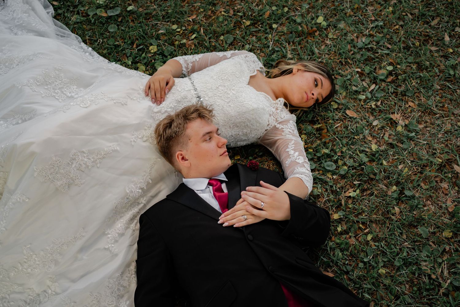 Bride and groom lying on grass in wedding attire, aerial view of couple in black suit and white lace gown.