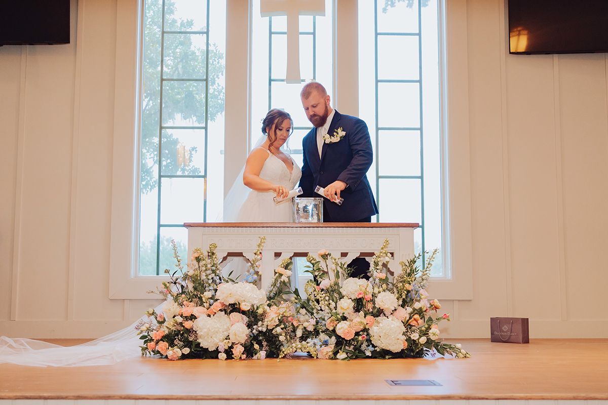 Bride and groom lighting a unity candle at a church altar adorned with white and pink floral arrangements.