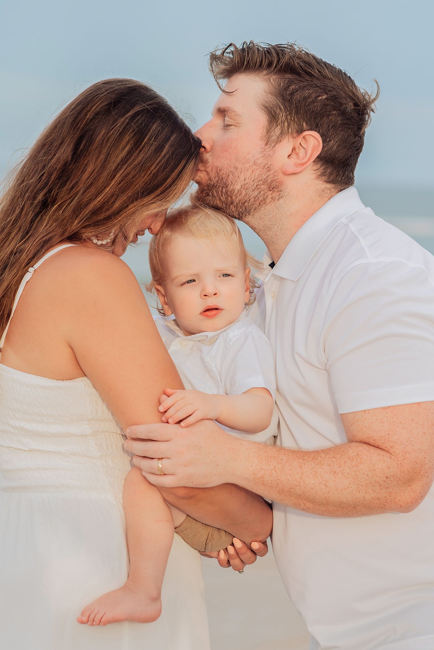 A couple shares a tender kiss while holding their baby in white outfits against a soft background in Jacksonville Florida on a beach