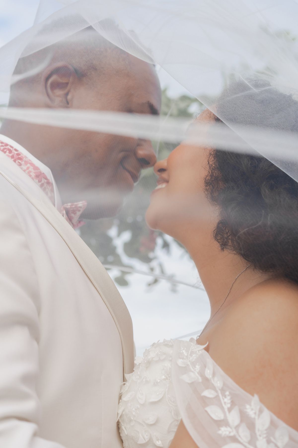 A bride and groom share a romantic moment under a wedding veil, nearly kissing in soft natural light.