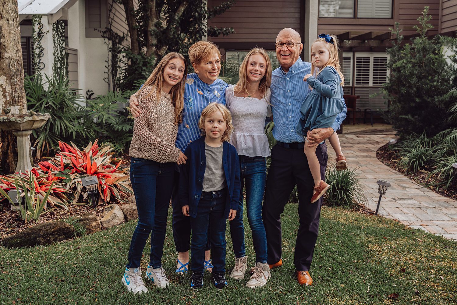 A group poses together outdoors in casual attire on a landscaped path with red plants in the garden for family photos in Jacksonville Fl.