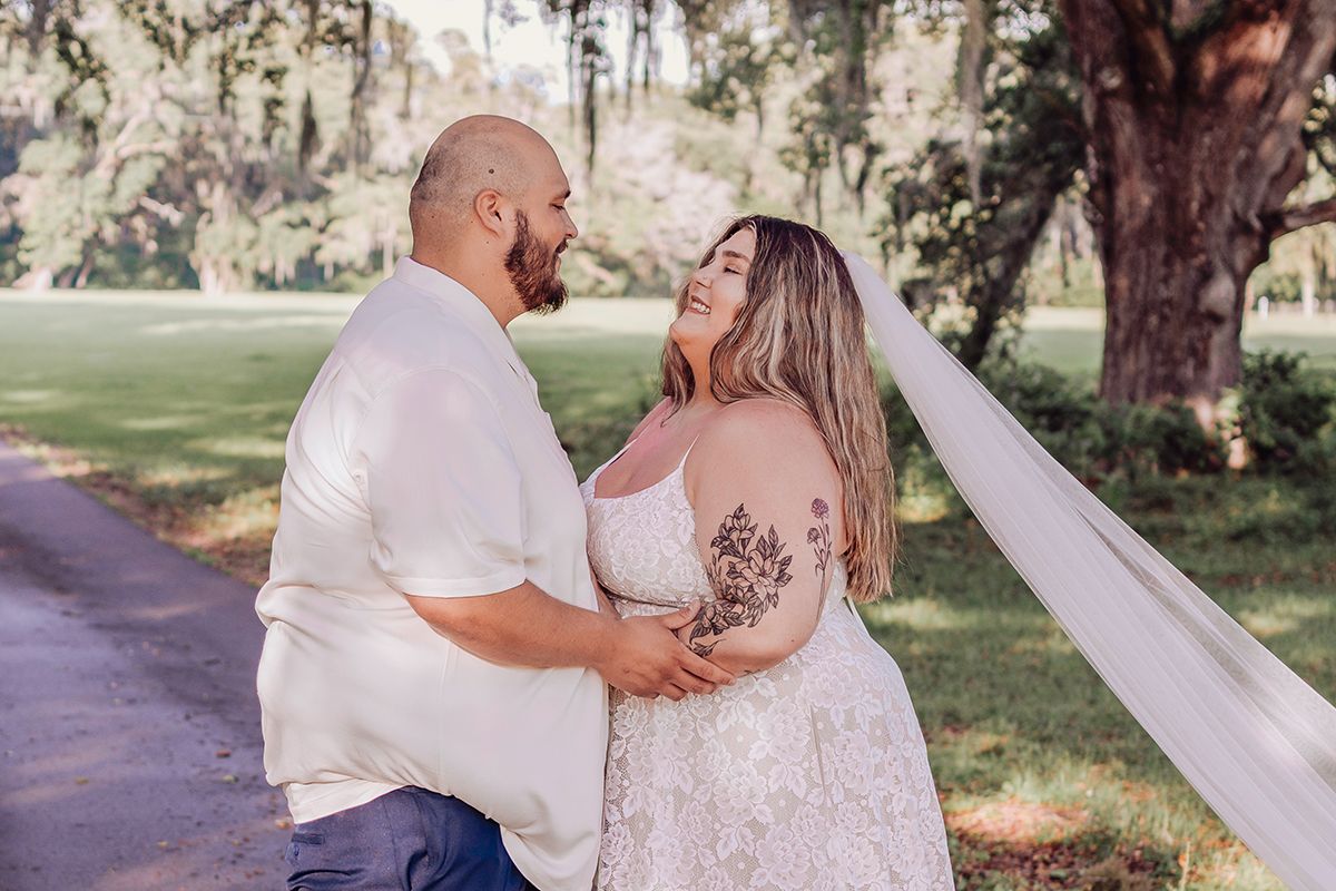 A couple in wedding attire embraces outdoors, bride in lace gown with veil, groom in white shirt, surrounded by trees.