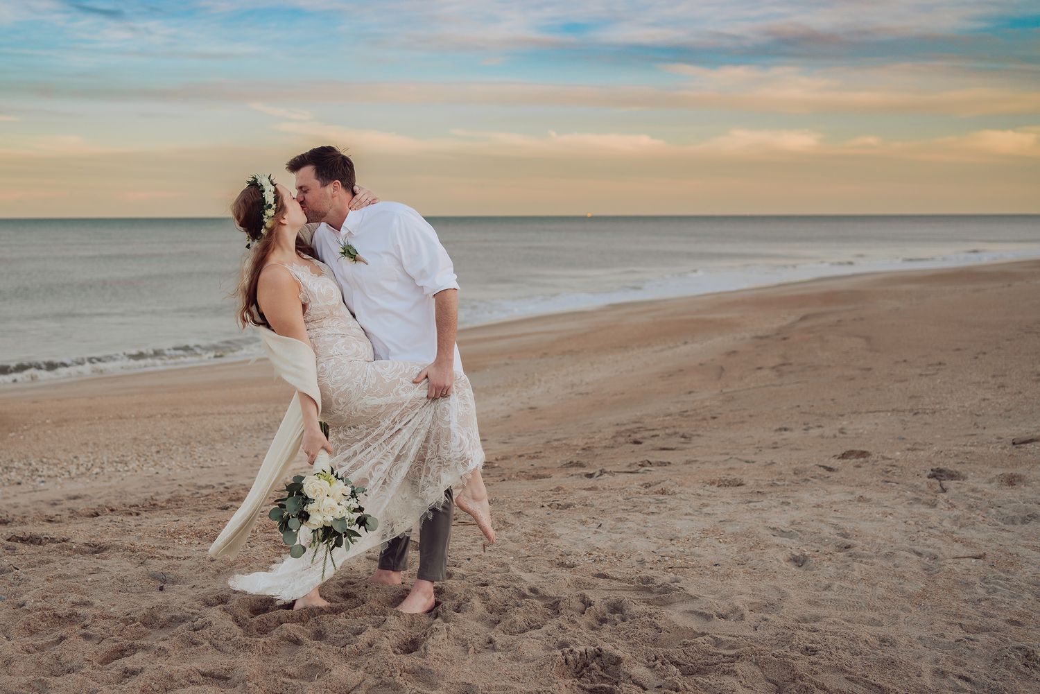 A couple shares a romantic moment on a sandy beach at sunset while holding a white floral bouquet.