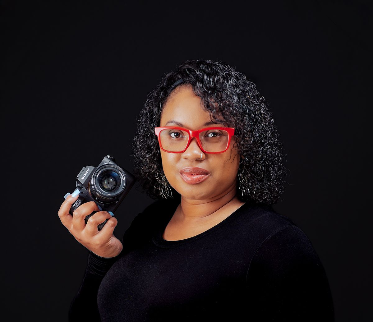 Professional photographer wearing red glasses and a black top holds a camera against dark background.