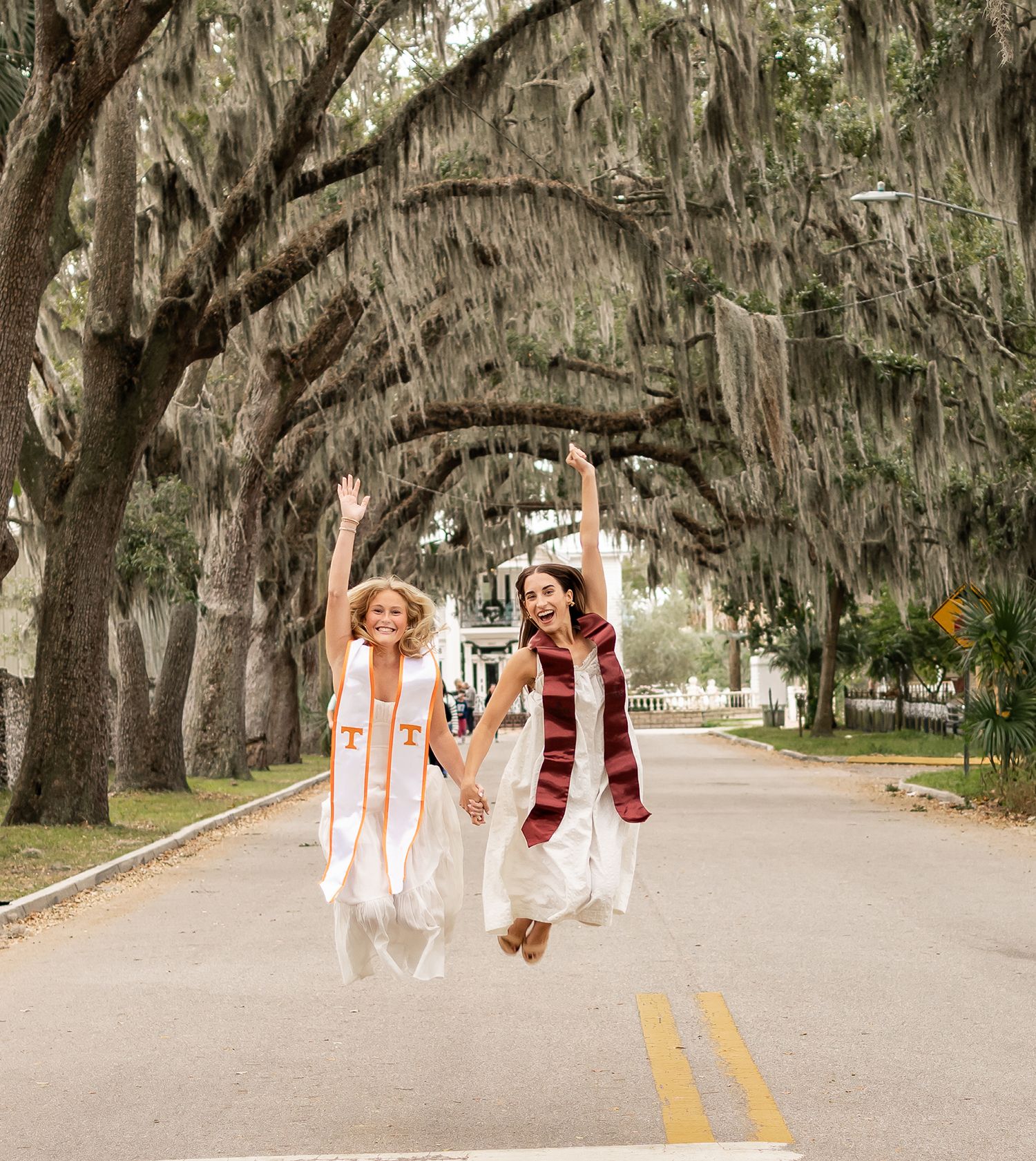 Two college graduates in white gowns and stoles joyfully jump on a tree-lined road celebrating their graduation day.