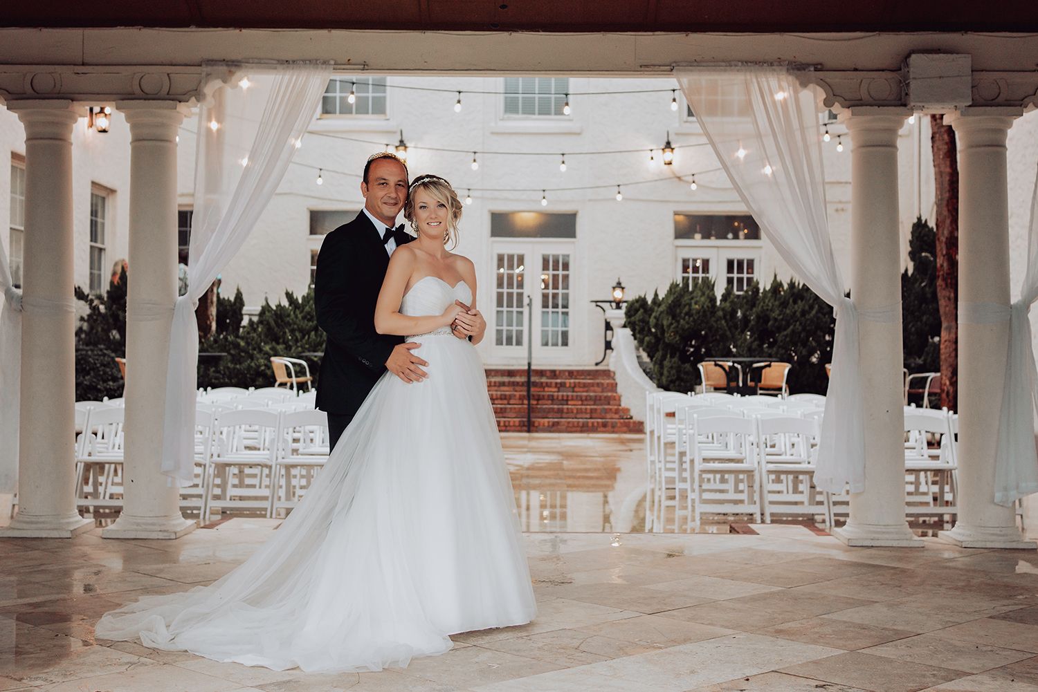 A couple dances together in a grand white ballroom with elegant columns and classic architectural details.